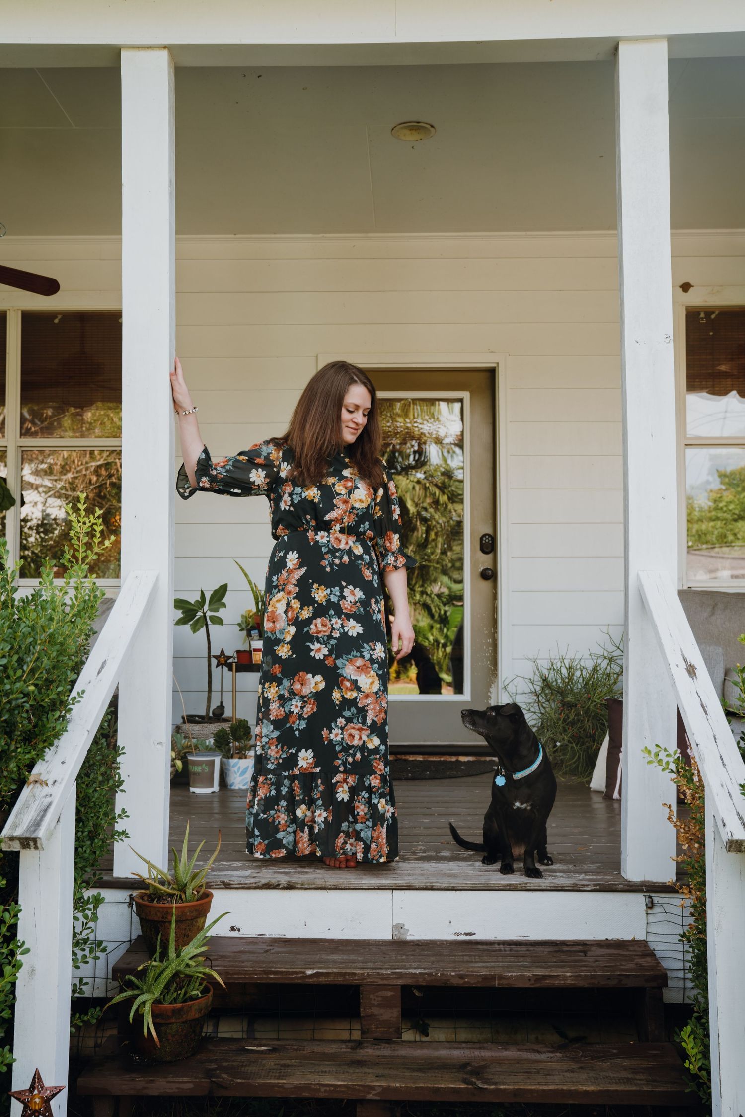 Woman in floral maxi dress standing on white farmhouse porch with black dog beside her.