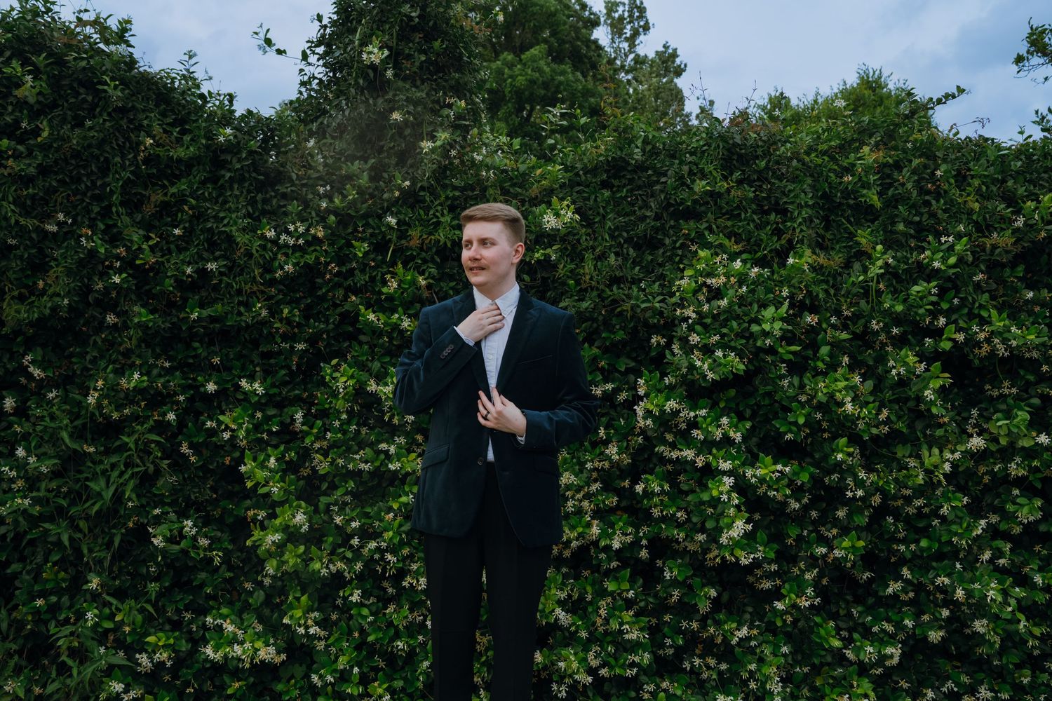 Young groom in dark suit adjusting tie while standing before tall green hedge outdoors.