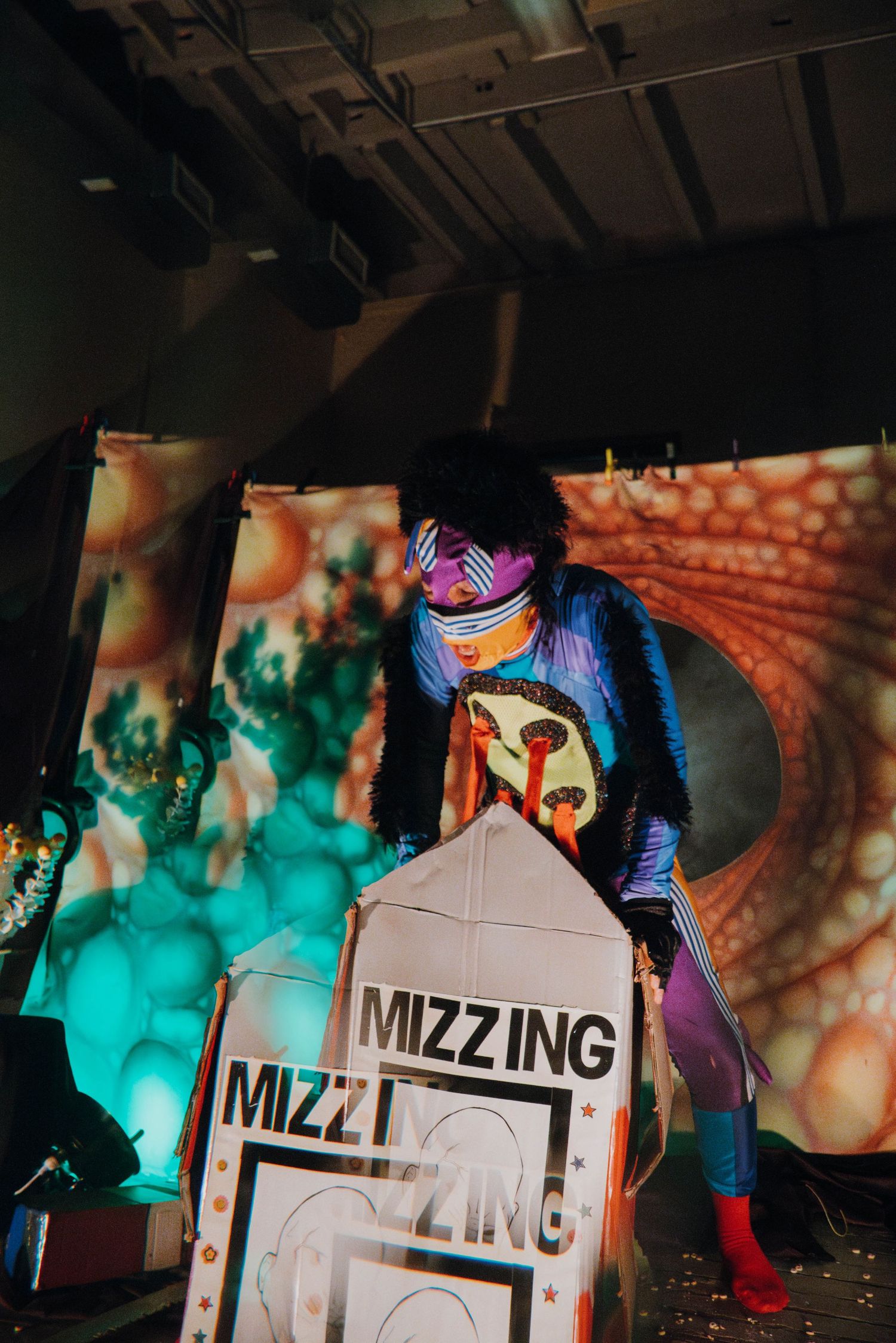 Colorful performer leans over Mizzing sign on eclectic illuminated performance stage.