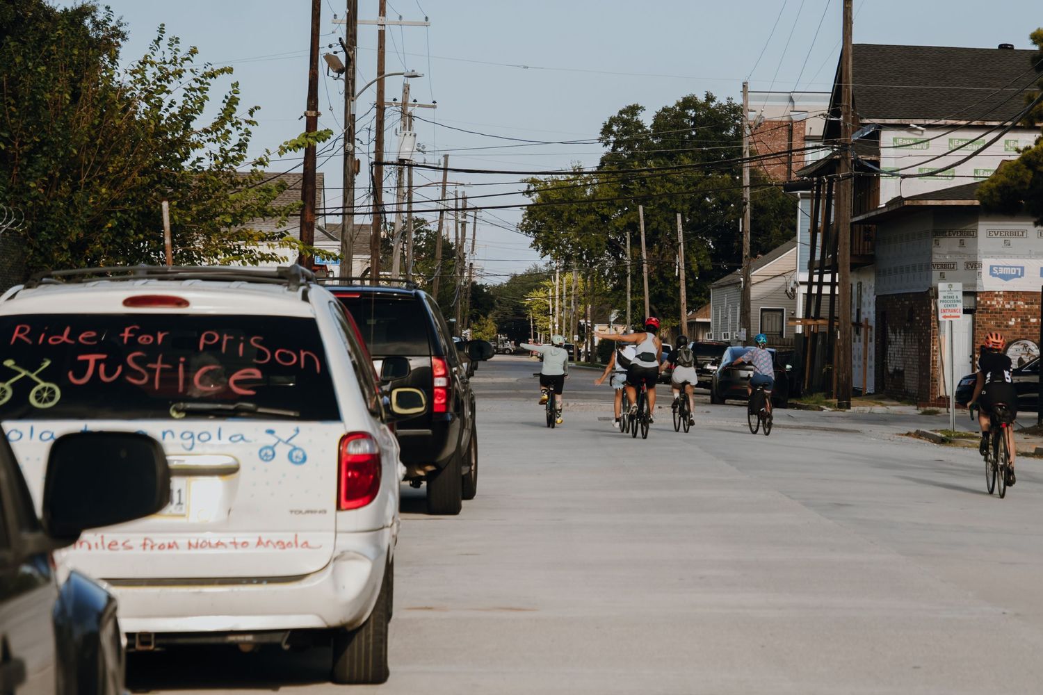 Cyclists riding down urban street past car with 'Ride for Prison Justice' sign displayed.