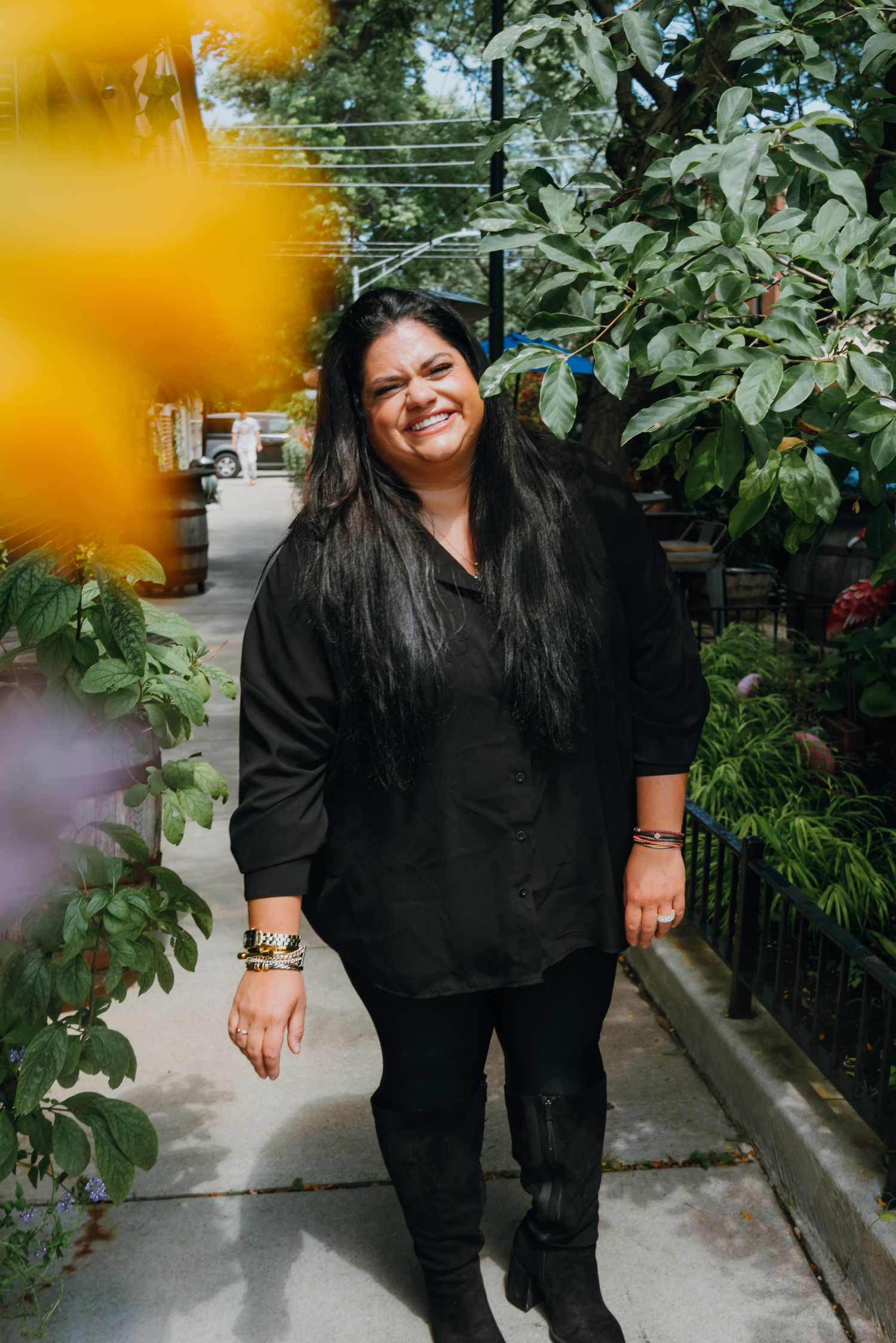Woman in all-black outfit smiling while walking along a flower-lined garden pathway on a cloudy day.