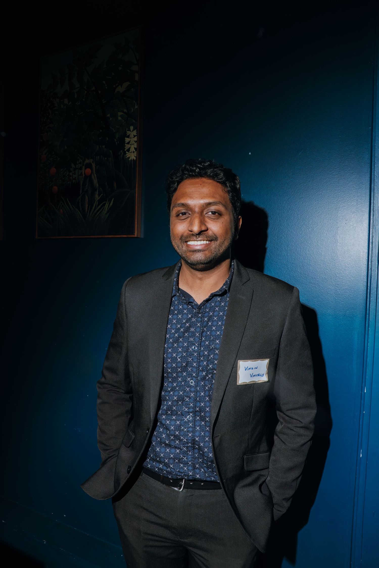 Smiling man in dark suit with name badge posing against teal blue wall at networking event.