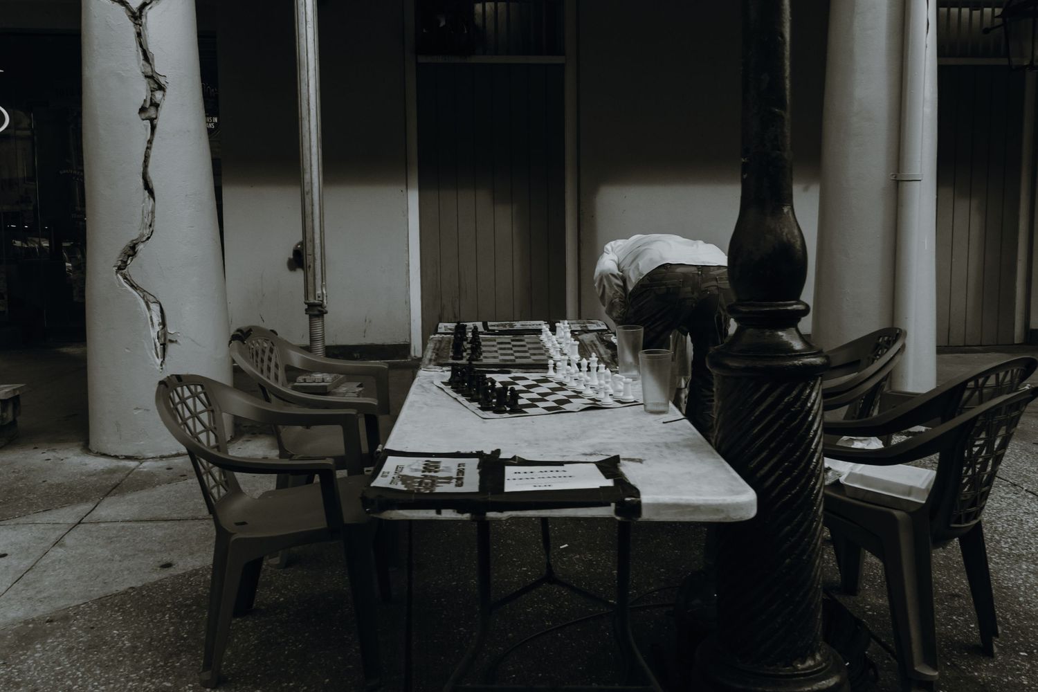 A person leaning over a chess board on a table in a dimly lit outdoor space.