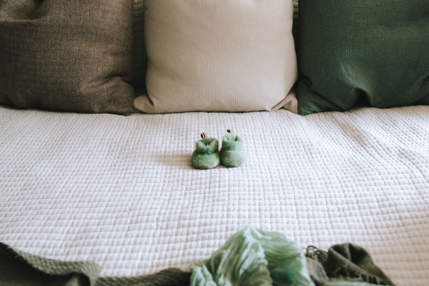 Tiny green frog baby shoes on a white quilted bed with green pillows.