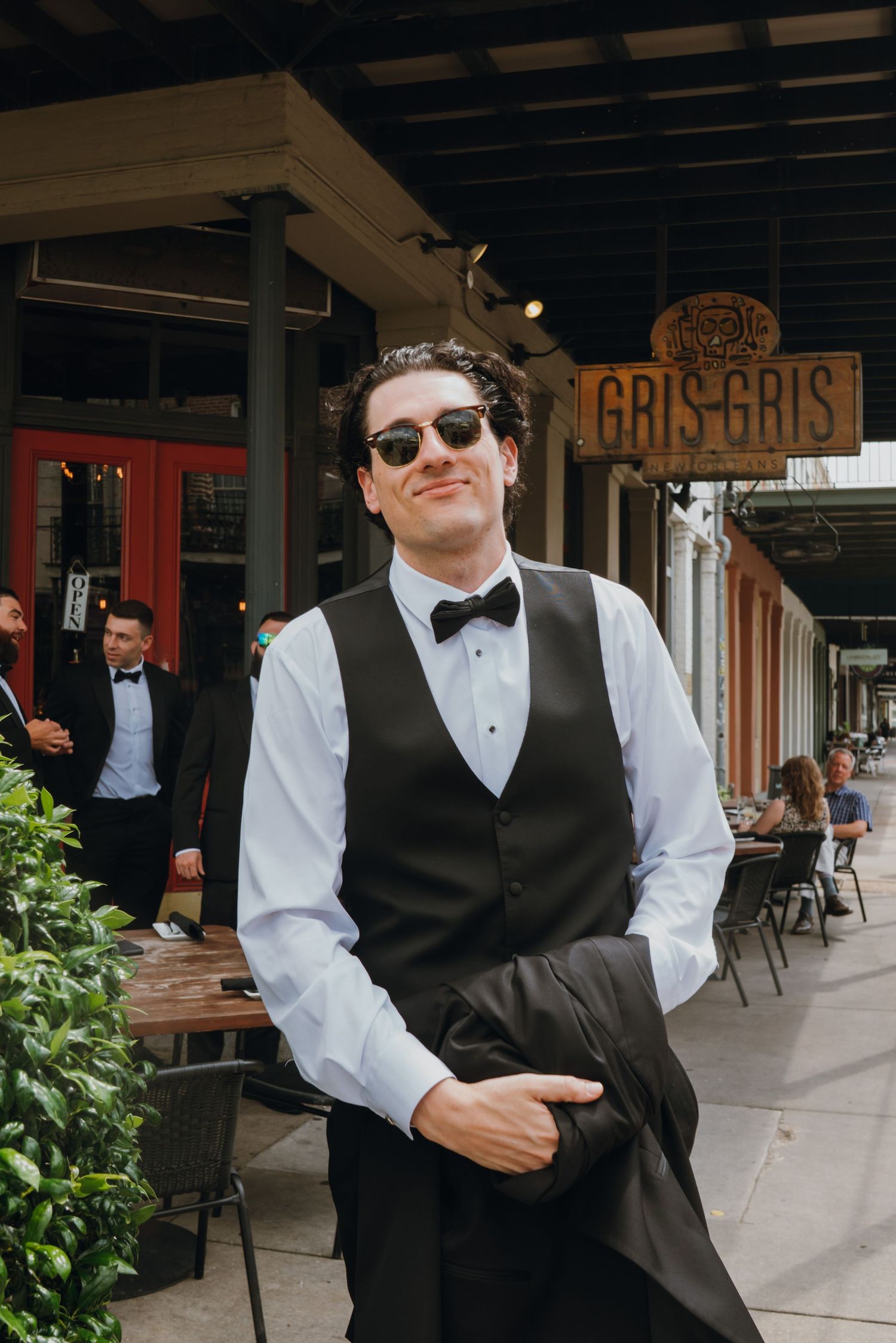 Stylish groom in black vest and bow tie with sunglasses standing outside Gris Gris restaurant.