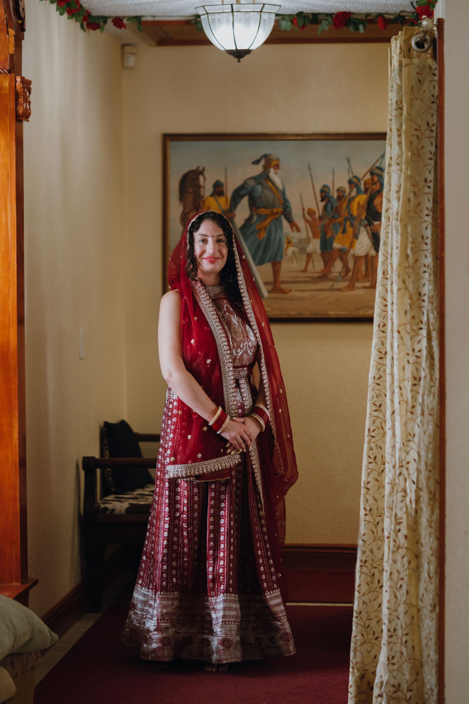 Smiling Sikh bride in ornate red and gold lehenga poses in a room with traditional Punjabi artwork.