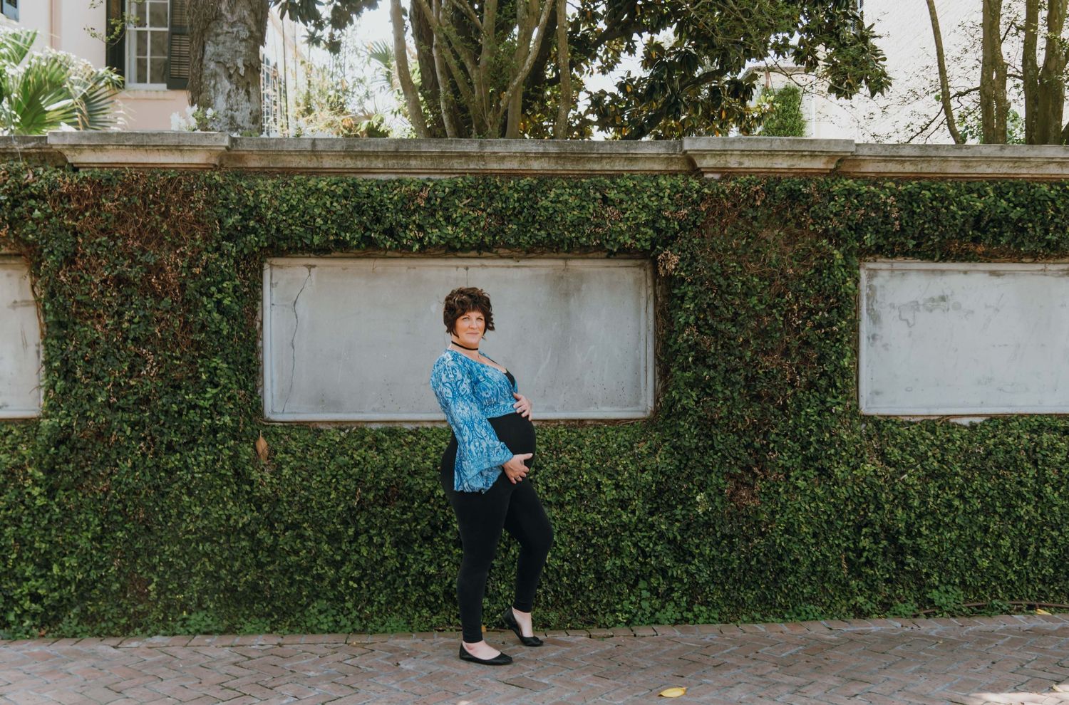 Pregnant woman in blue patterned top standing against ivy-covered wall during maternity photoshoot.New Orleans, Louisiana, Mississippi