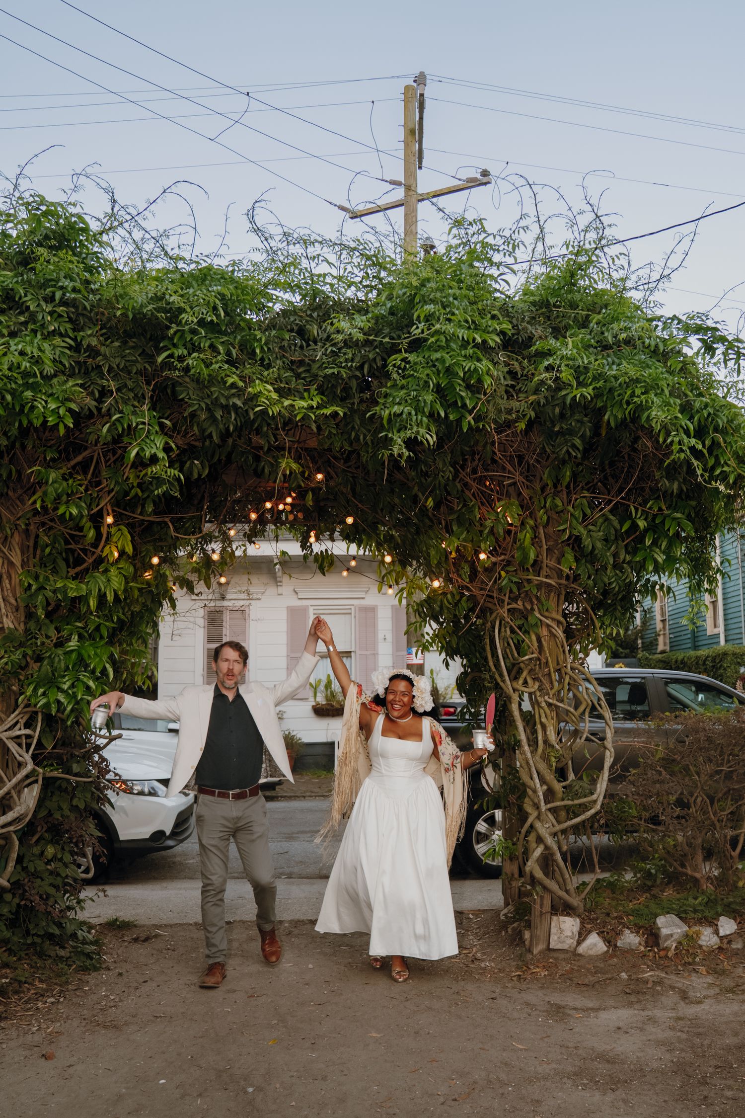 Joyful newlyweds celebrate under a vine-covered arch adorned with string lights at dusk.