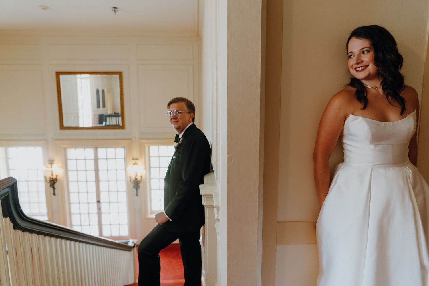 Bride in strapless white dress smiling as groom approaches up a red-carpeted staircase for first look. New Orleans, Louisiana, Mississippi