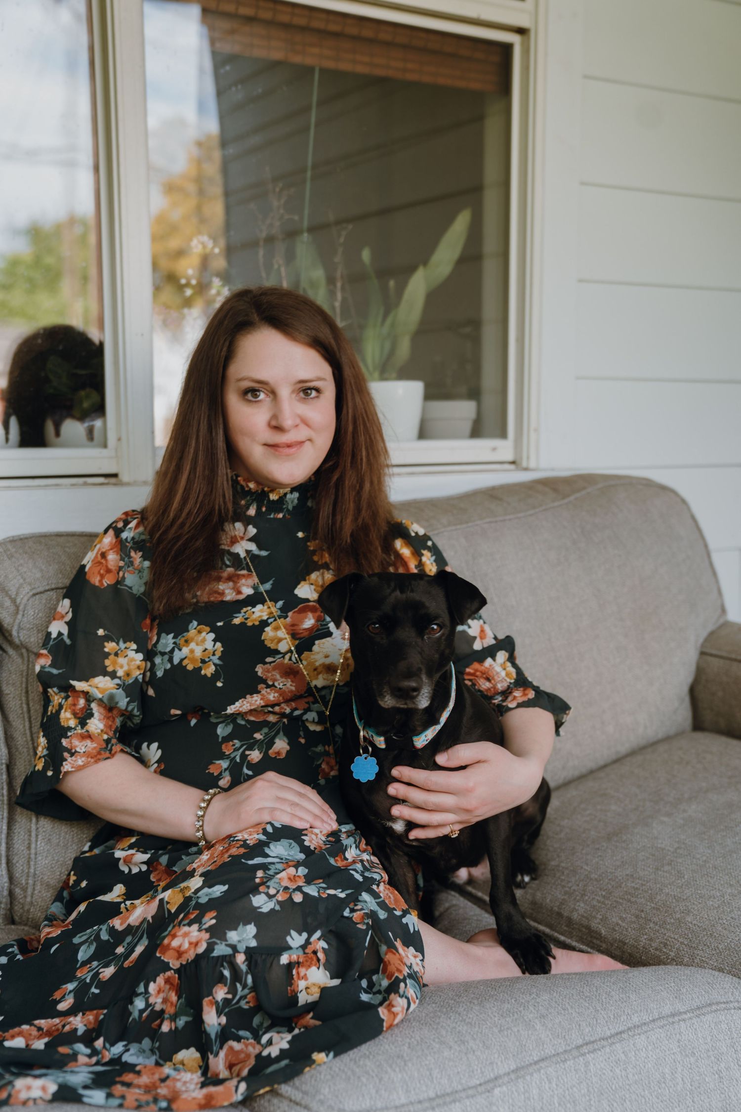 Woman in floral dress sitting on porch sofa with black dog wearing blue tag.