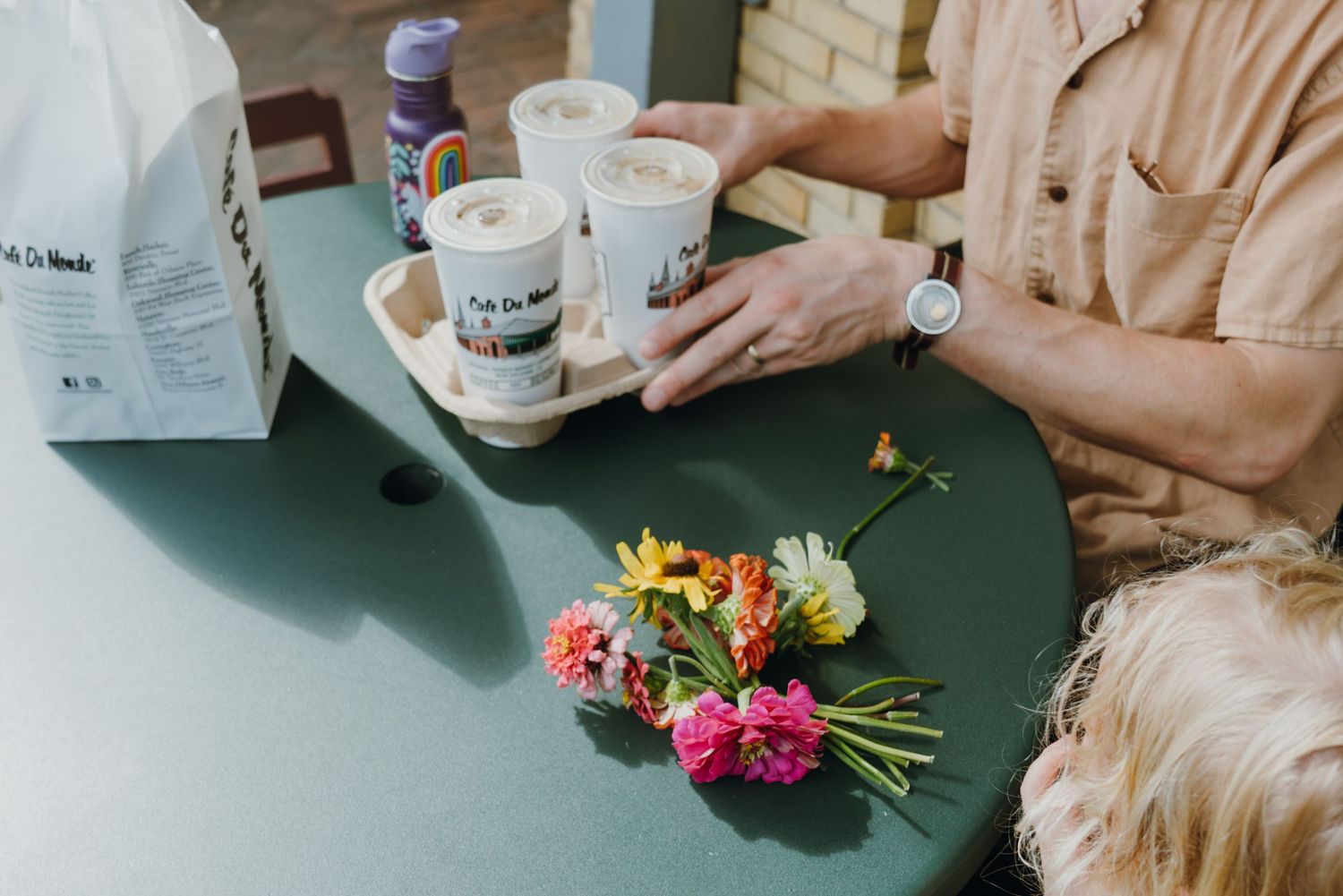 Person at green table with coffee cups in carrier and colorful flower bouquet, casual outdoor cafe setting.