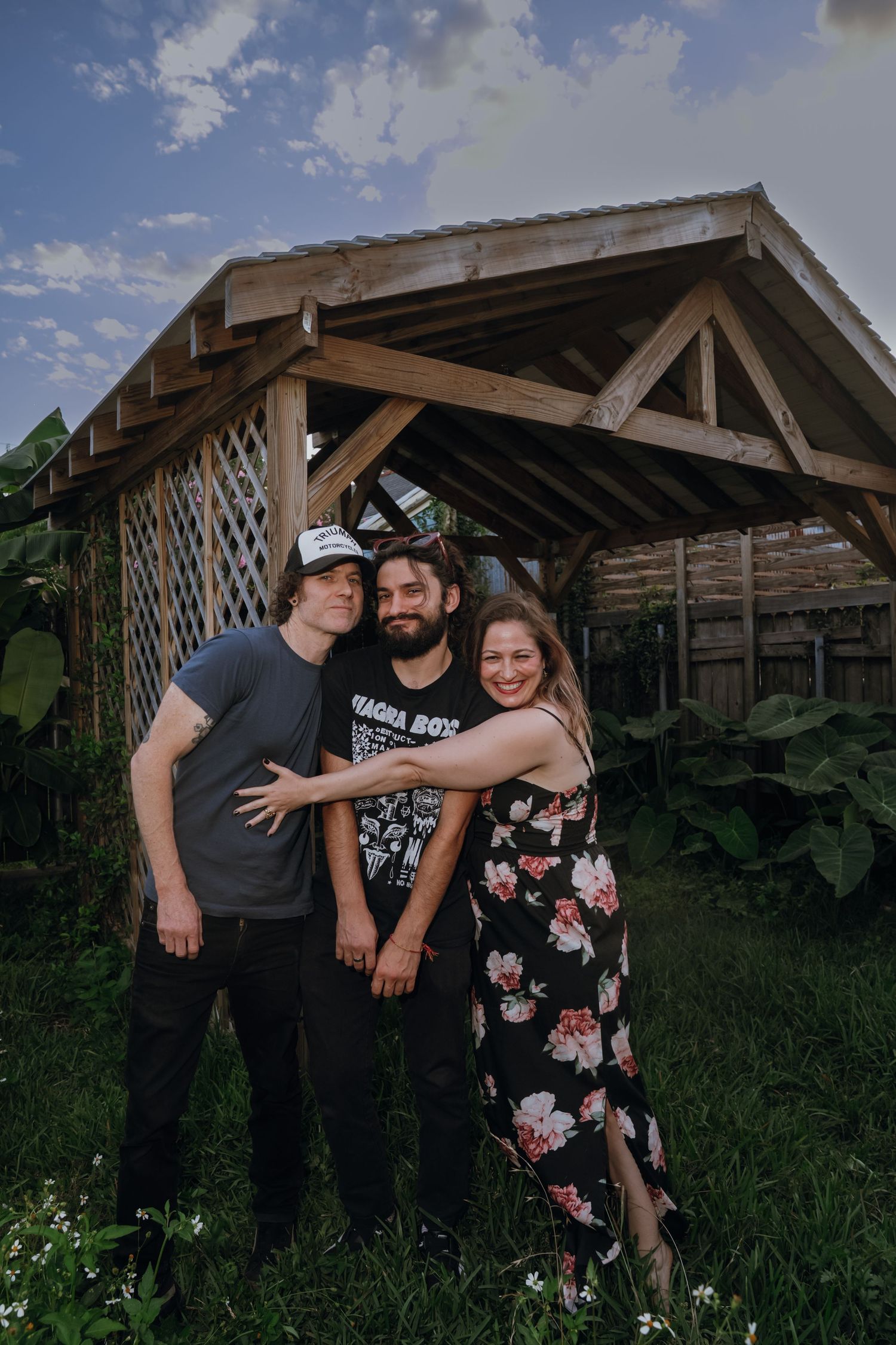 Three friends smiling and posing together in a lush backyard garden beneath a rustic wooden pergola.