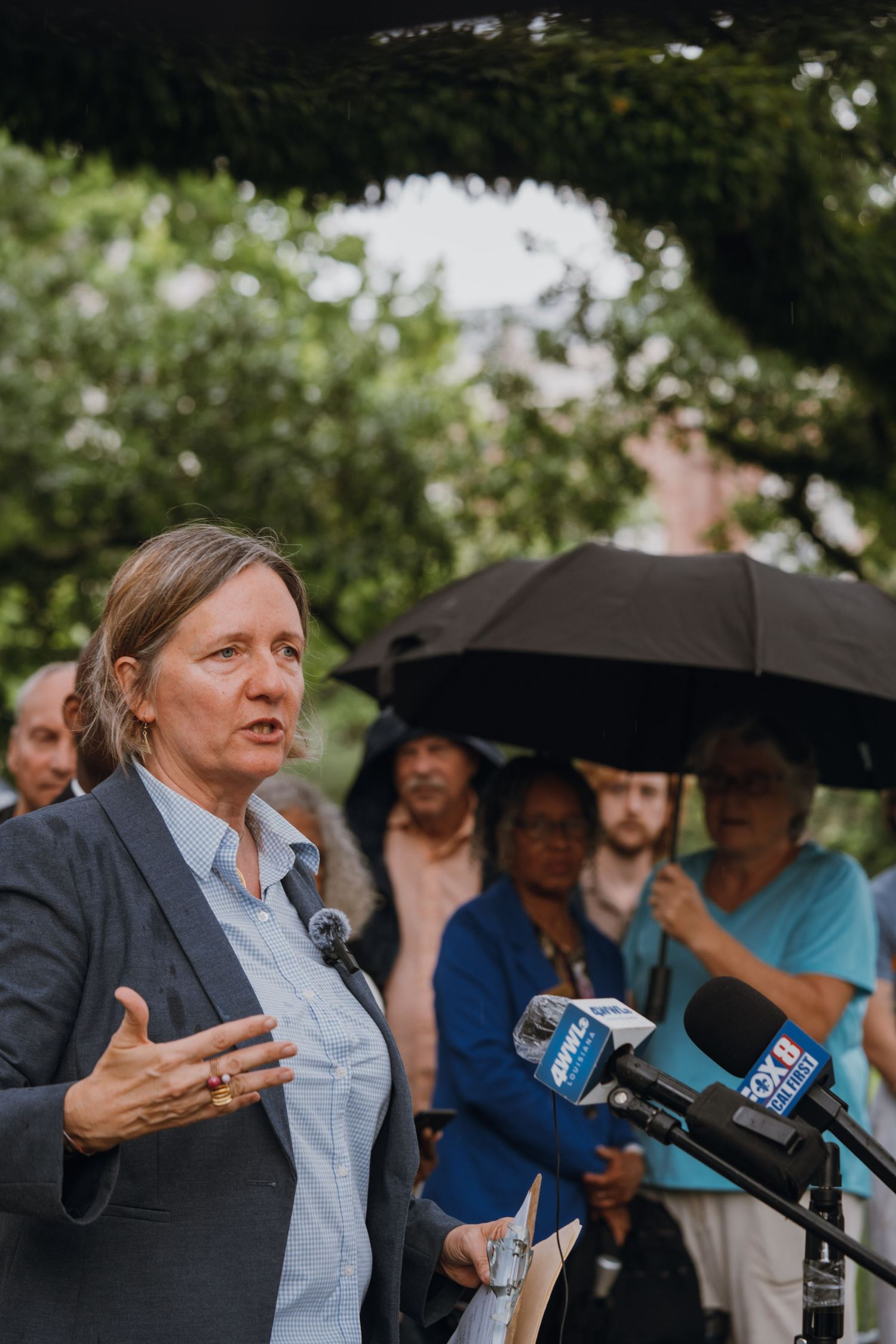 Woman speaking at an outdoor press conference with microphones, gesturing while addressing the media.