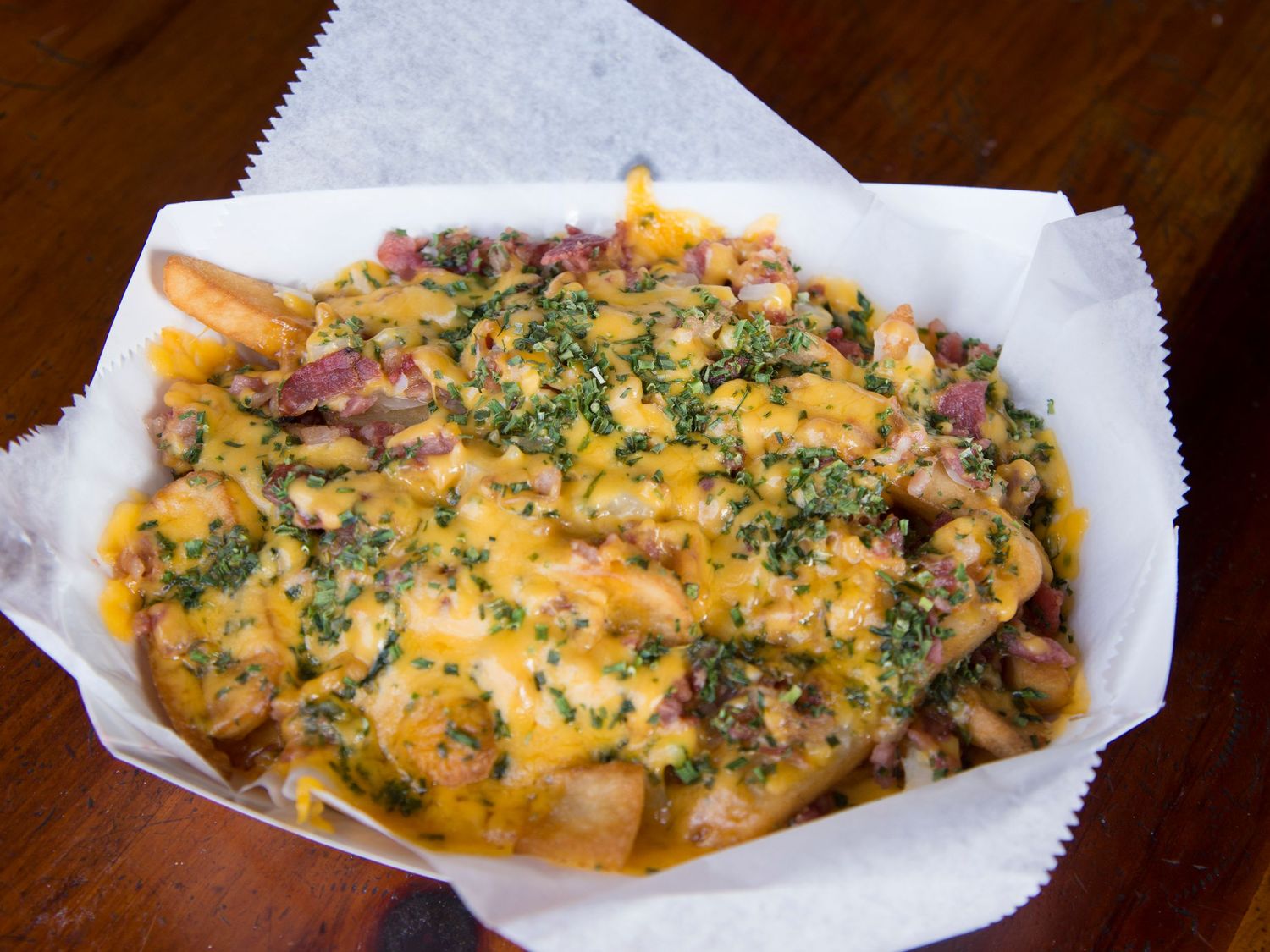 Loaded cheese fries topped with melted cheddar, bacon, and fresh herbs served in a paper-lined basket.
