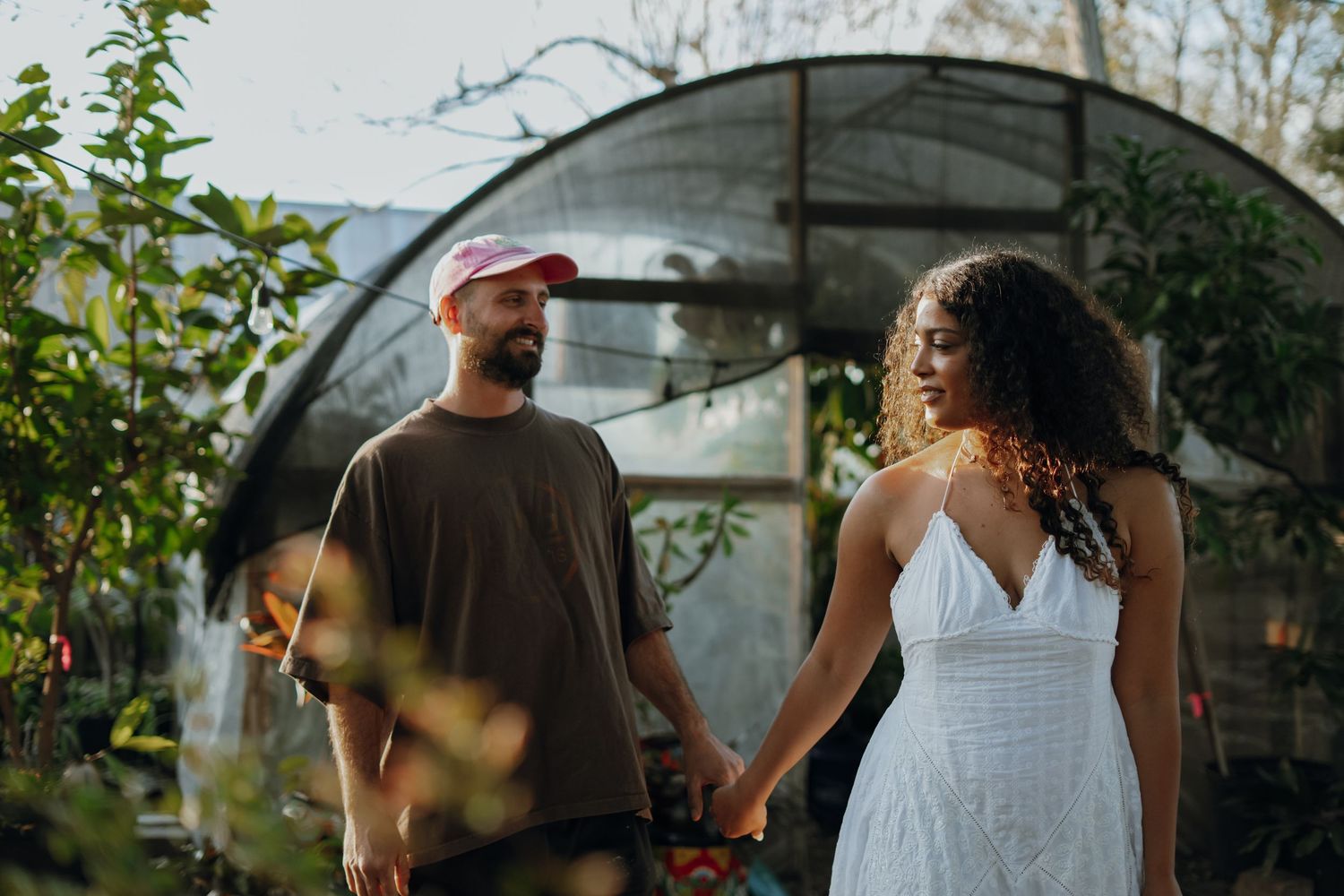 An interracial couple holds hands and walks near a greenhouse surrounded by lush green plants at sunset.