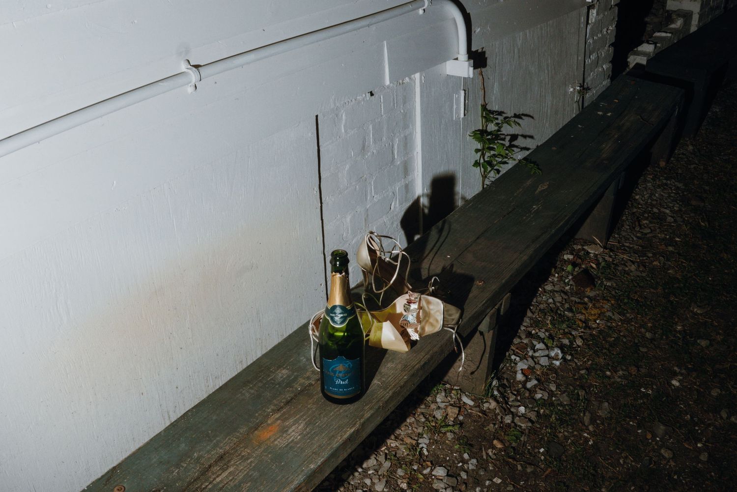 Champagne bottle and glasses left on wooden bench against white wall at wedding venue.