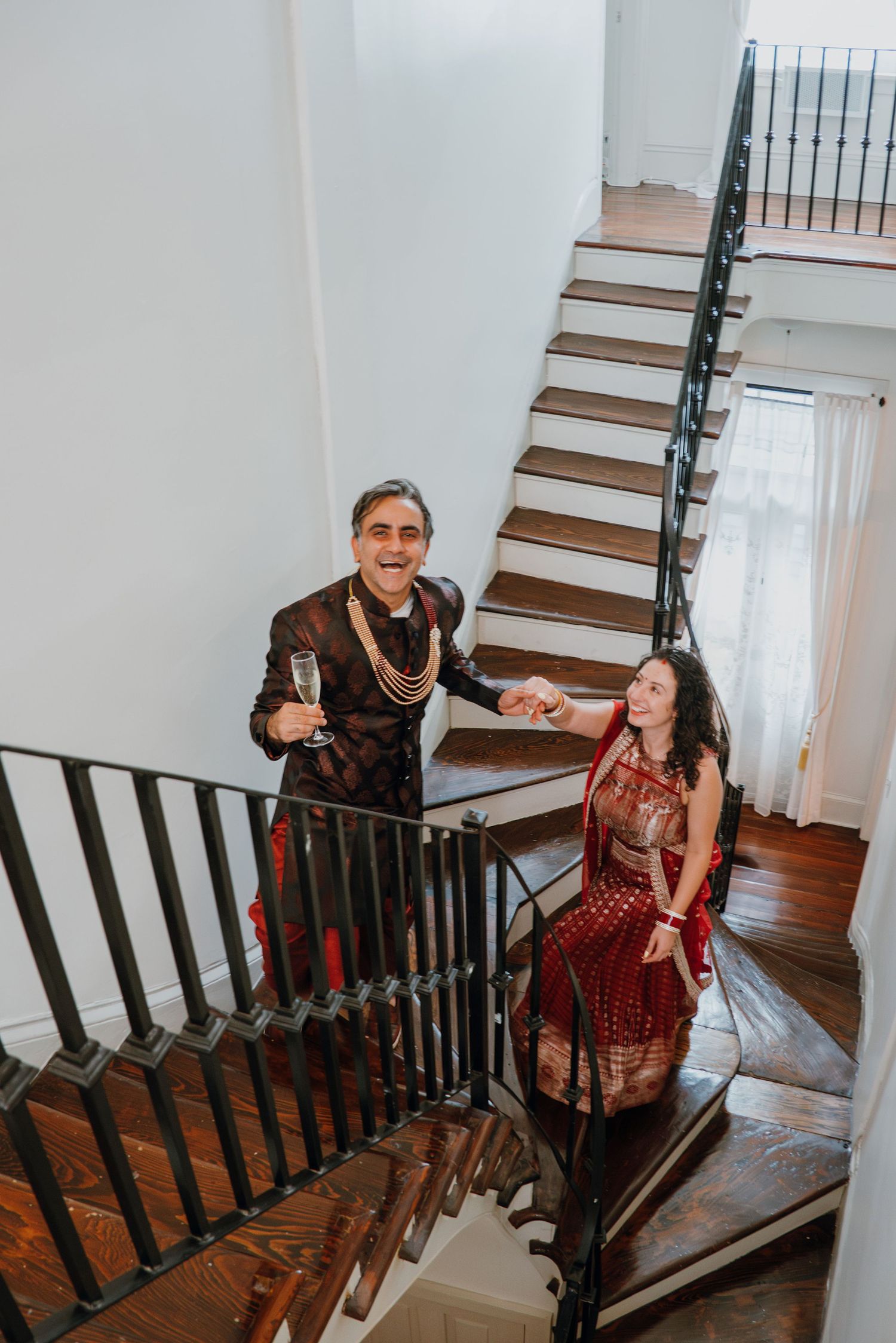 Couple in traditional Indian wedding attire descending an elegant staircase, man holding champagne glass.
