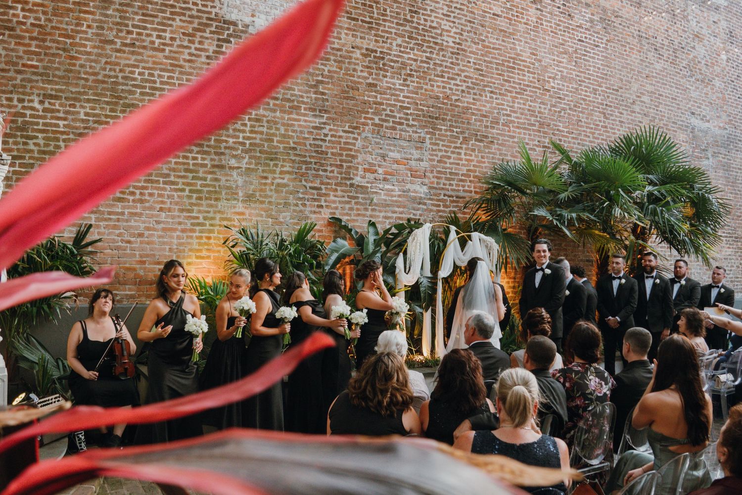 Outdoor wedding ceremony against brick wall with bridesmaids in black dresses, tropical plants, and decorative arch.