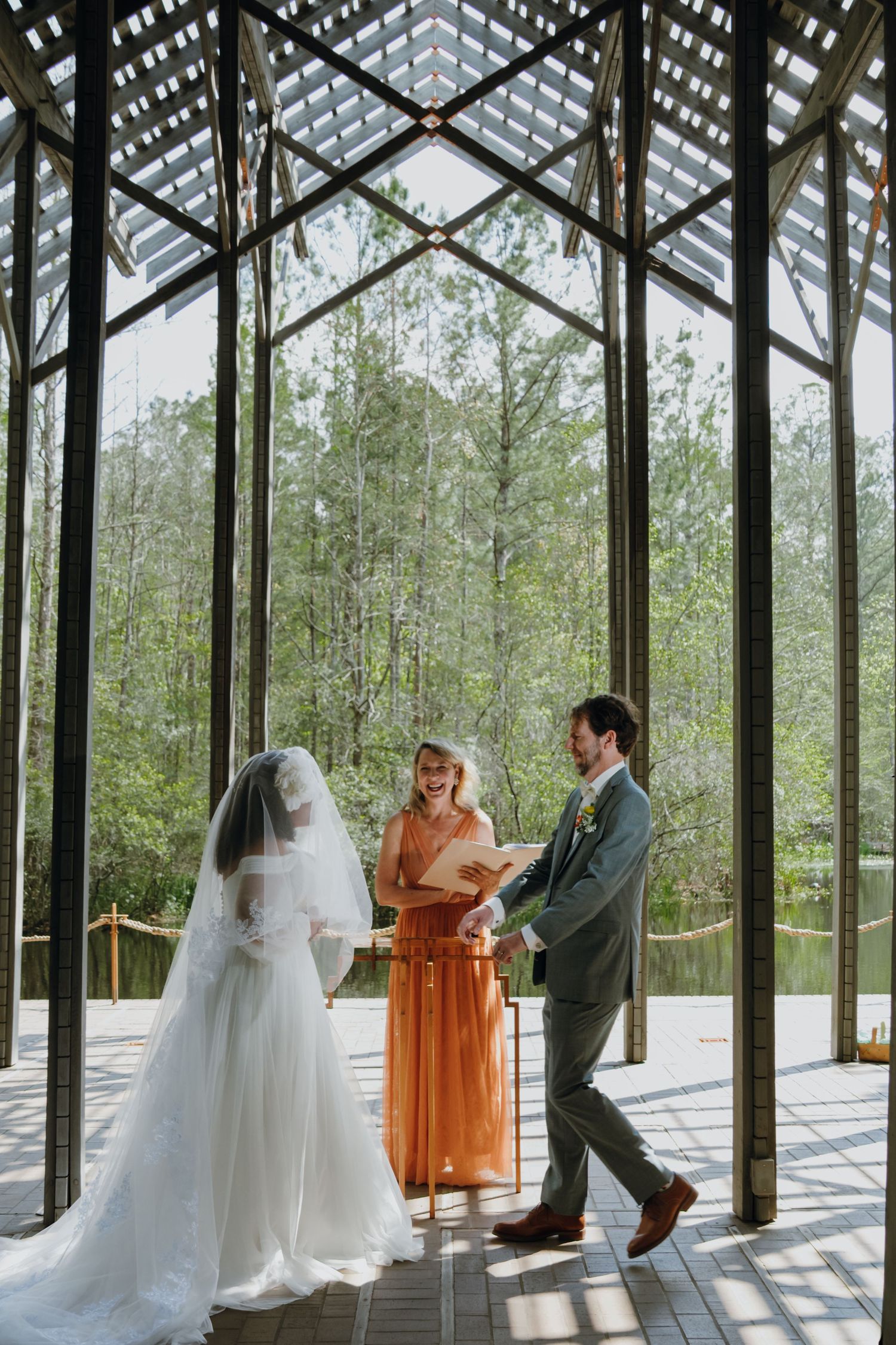 Couple laughs with officiant during wedding ceremony in stunning glass chapel.