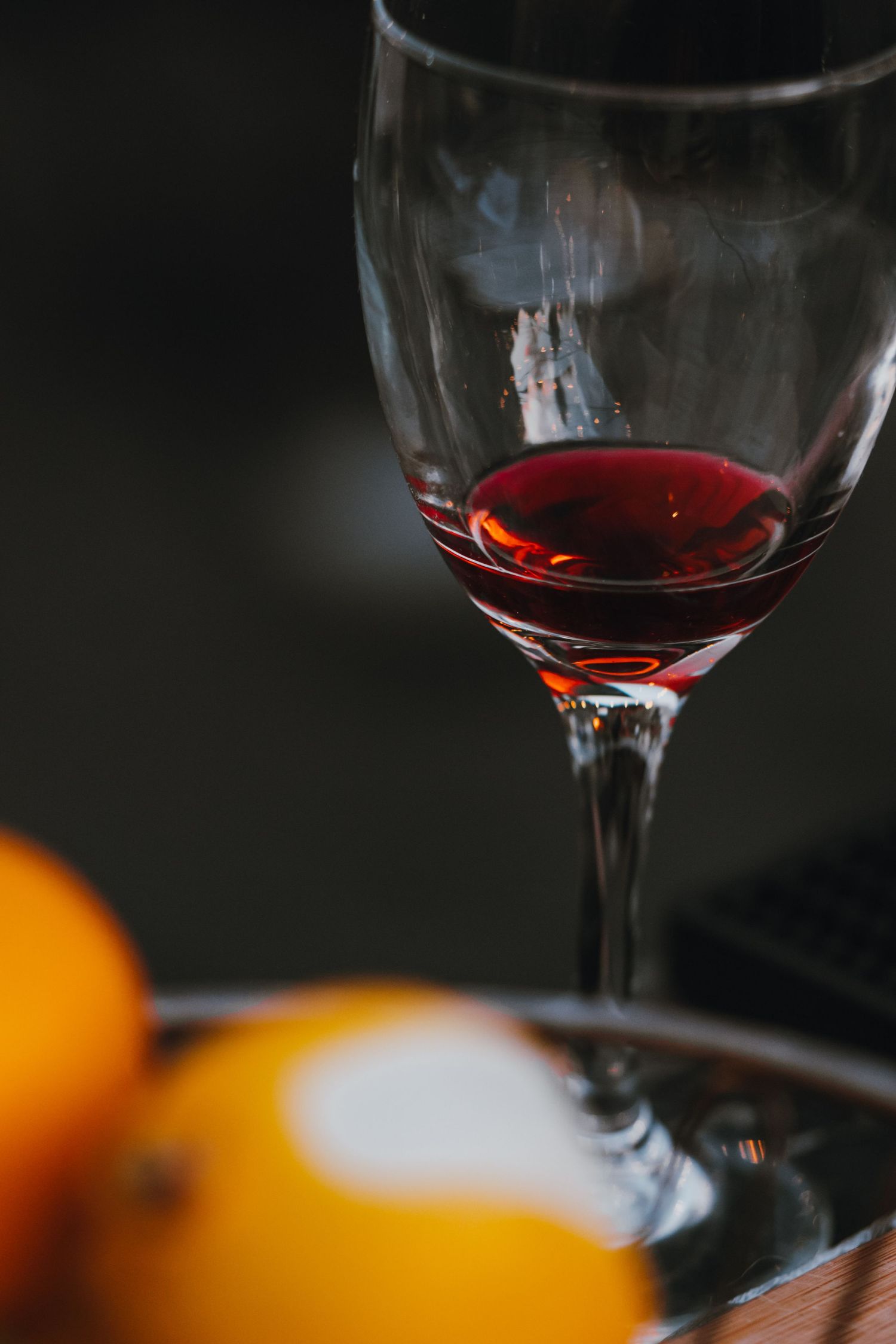 A wine glass with red wine on a dark background with blurred yellow fruit in the foreground.