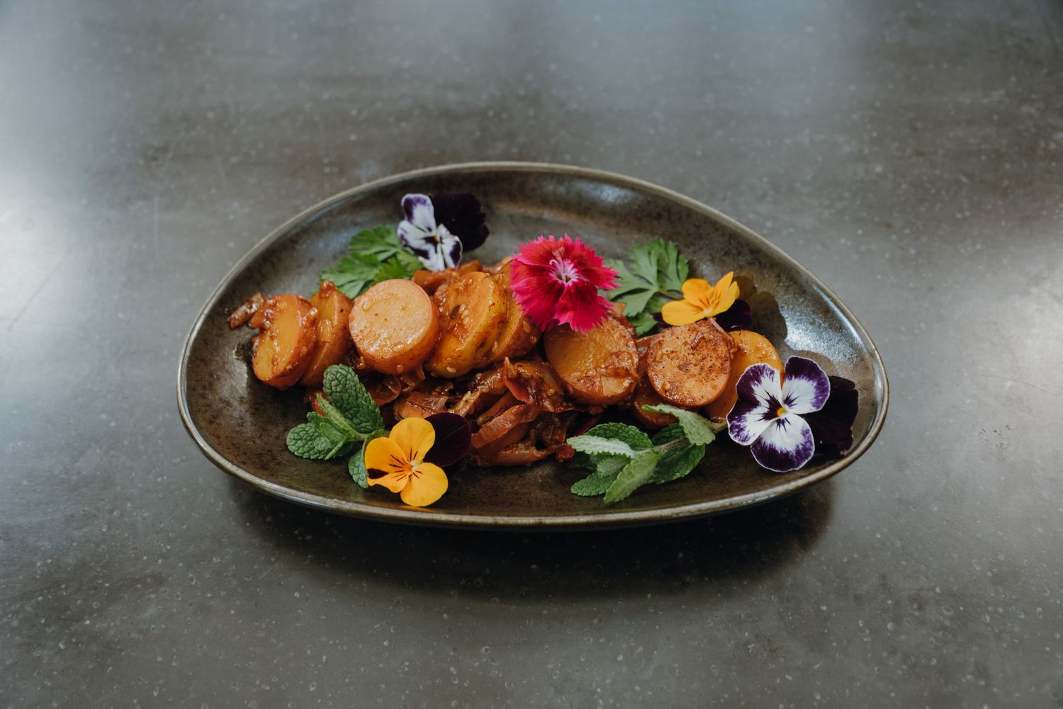 Elegant oval dark ceramic plate with roasted vegetables, fresh greens, and colorful edible flowers on gray surface.