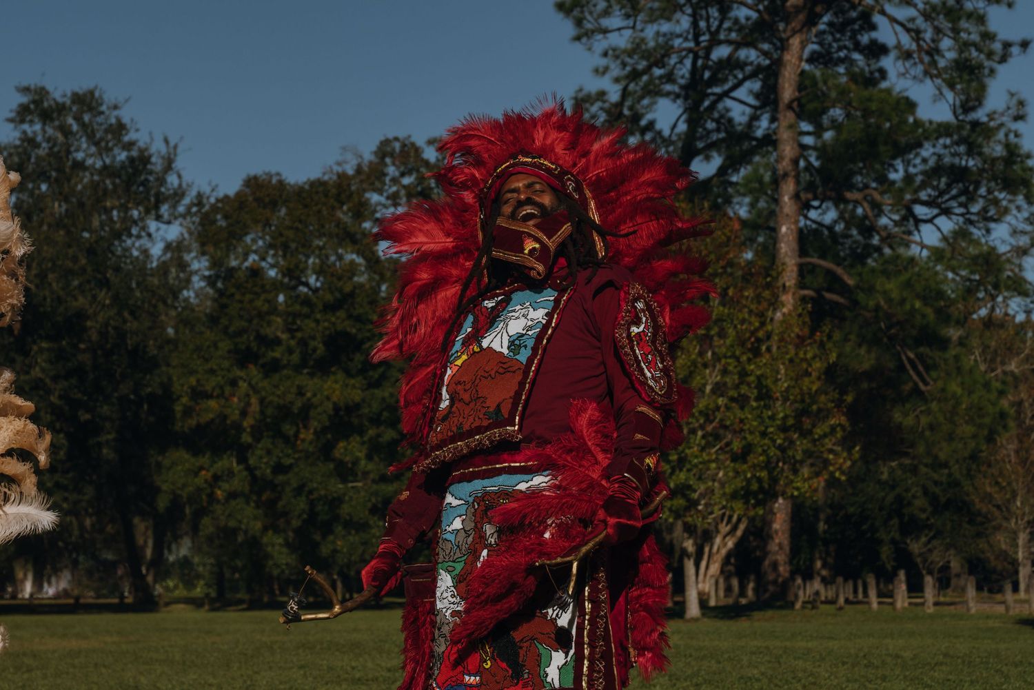 Mardi Gras Indian in striking all-red feathered headdress and beaded suit stands on grass in sunlight.