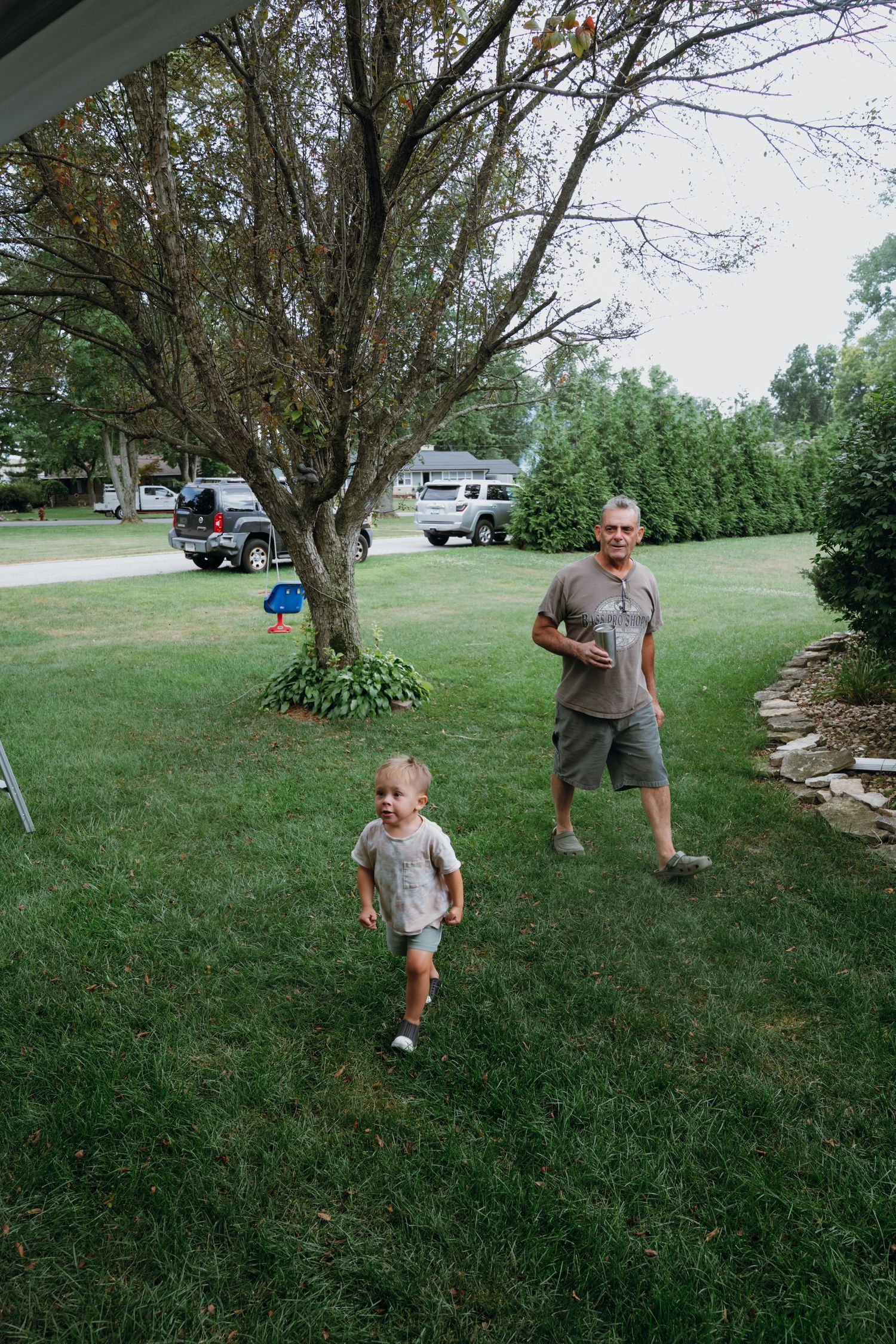 Grandfather and toddler walking together on a green lawn outside a suburban home.