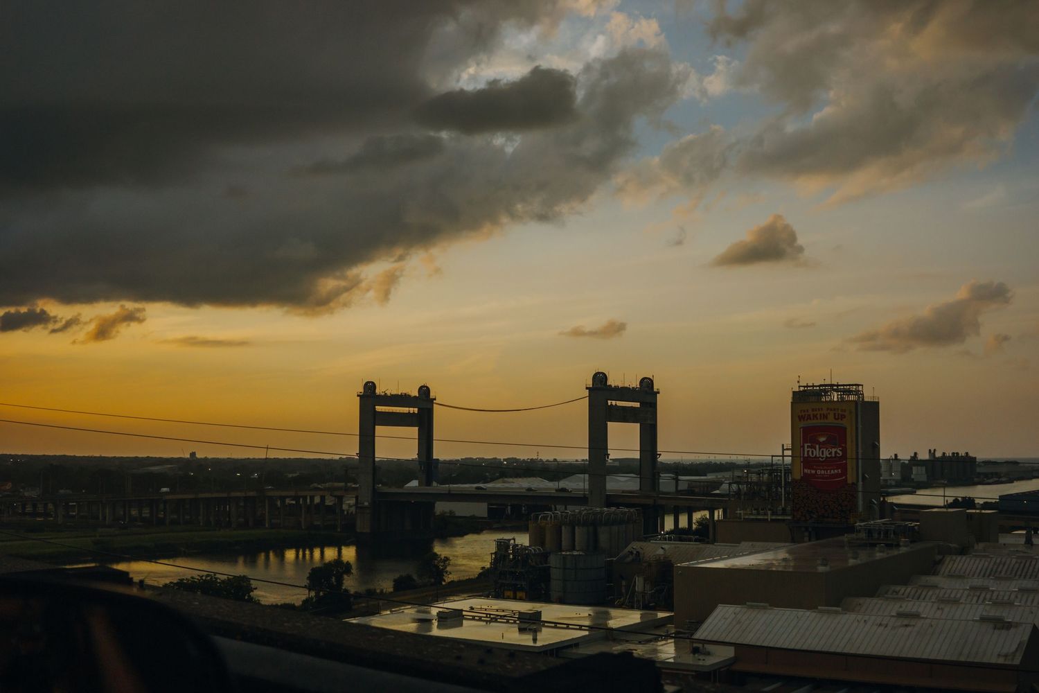 Dramatic sunset over a city bridge with dark storm clouds and golden sky reflecting on water.
