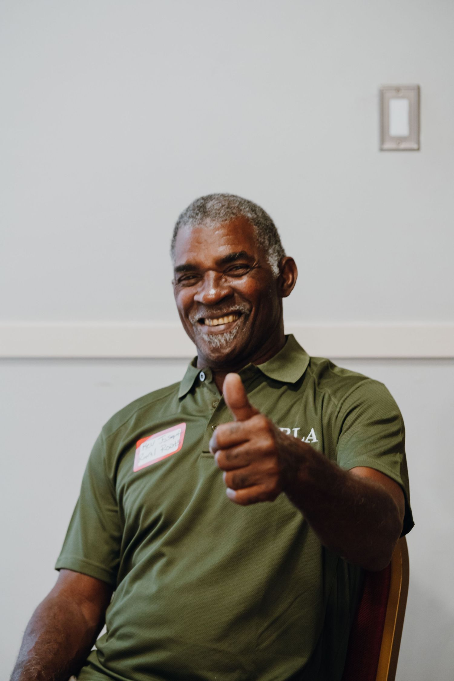 Smiling man in green polo shirt giving thumbs up while wearing name tag at community event.