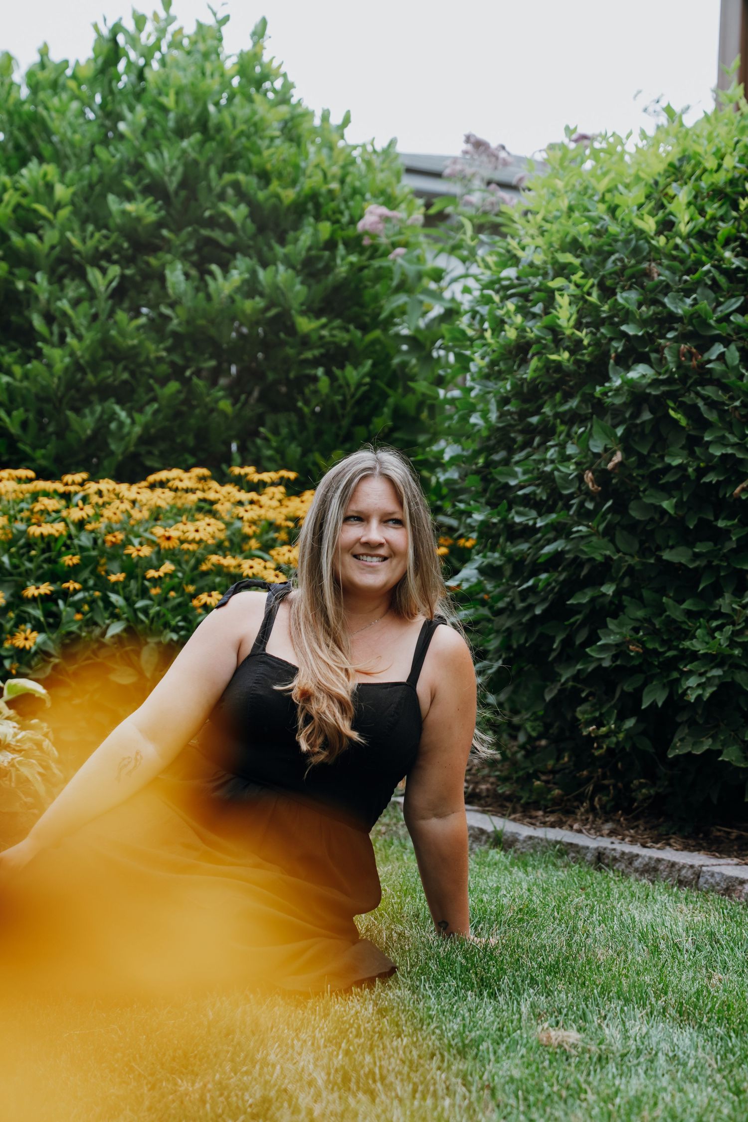A smiling blonde woman in a black dress poses in a lush garden with yellow flowers and green hedges.