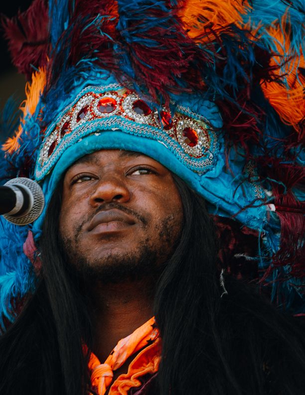 Man wearing vibrant blue and orange feathered Mardi Gras Indian headdress with red jewel decorations performing at outdoor event.