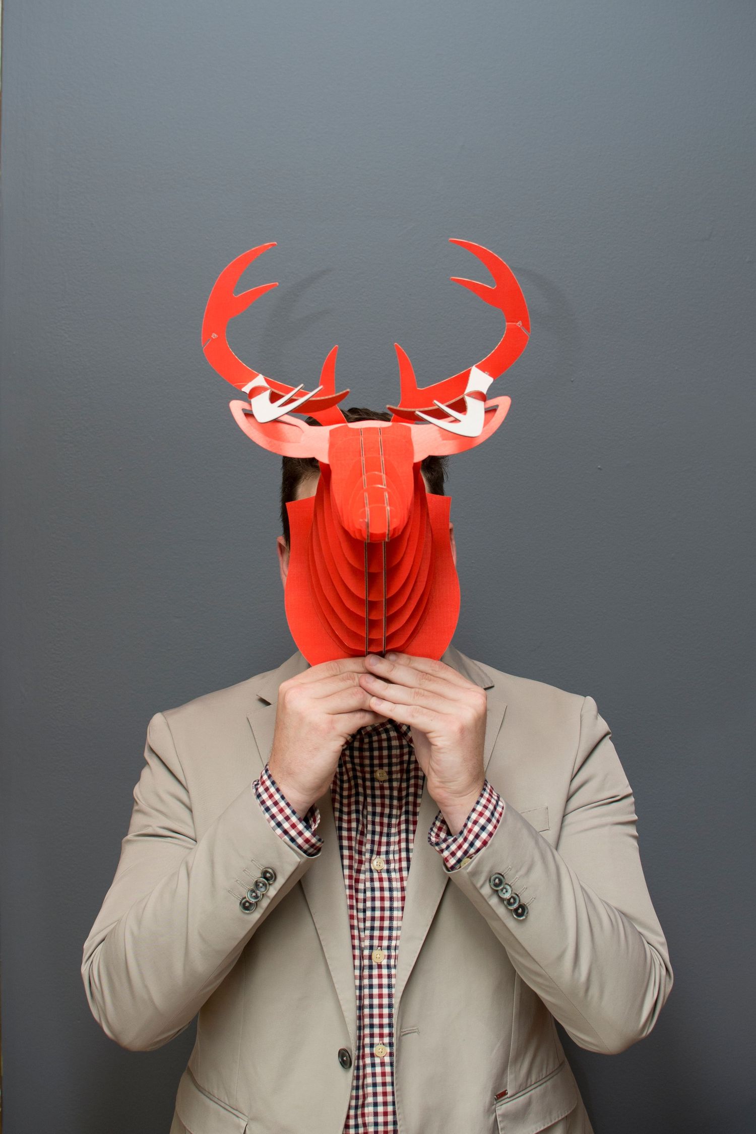 Man in beige blazer holding a red paper deer mask in front of his face against gray background.