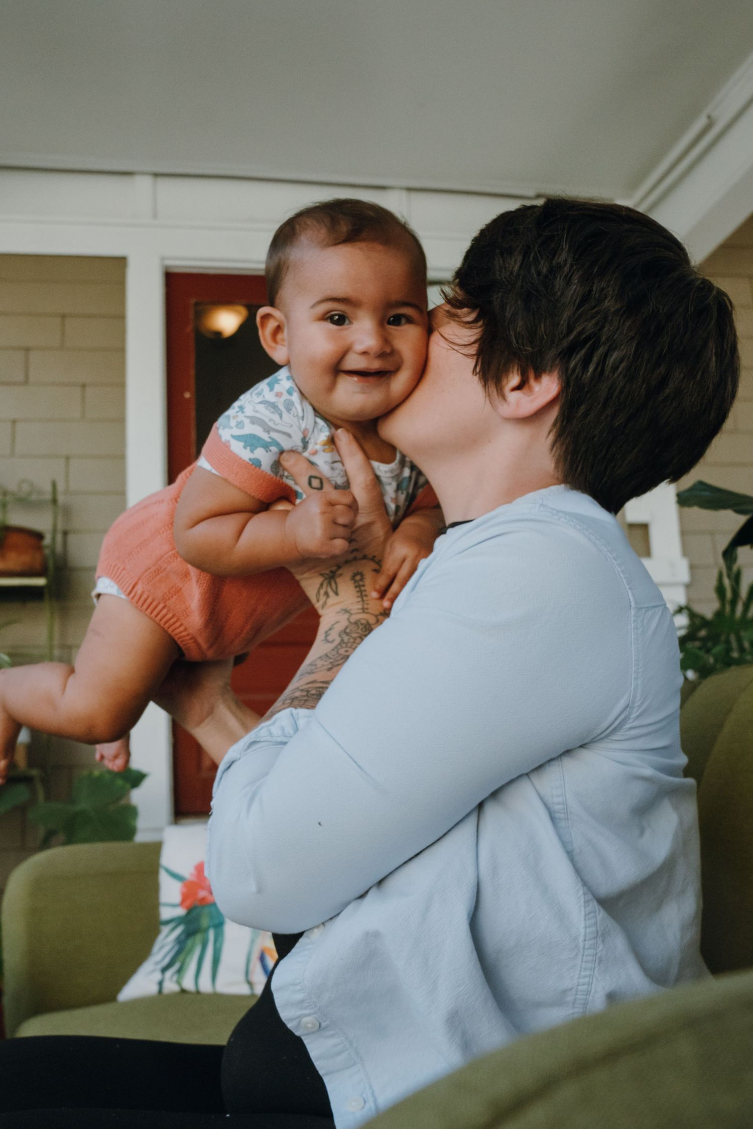 Parent kissing a smiling happy baby on the cheek while holding them up indoors near a porch.