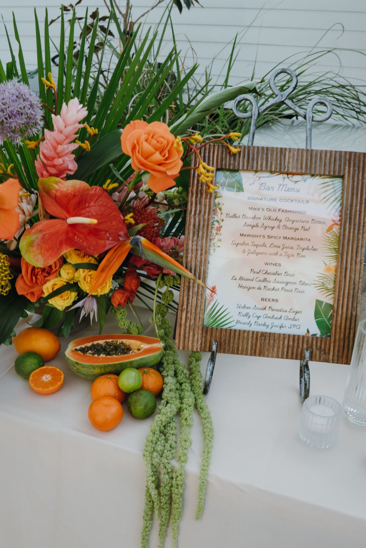 Tropical wedding bar menu display with vibrant orange roses, red anthuriums, and fresh papaya and citrus fruits on a white table.