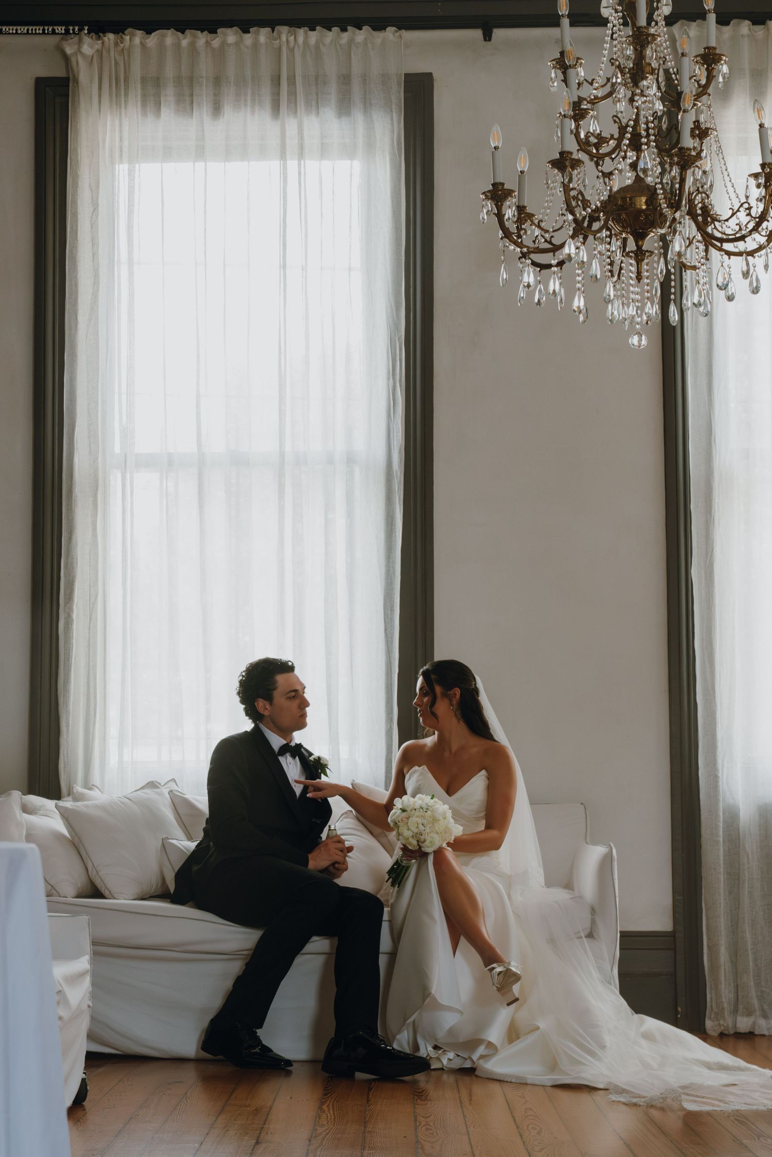 Bride and groom sharing a romantic moment on white sofa beneath crystal chandelier in elegant wedding venue.