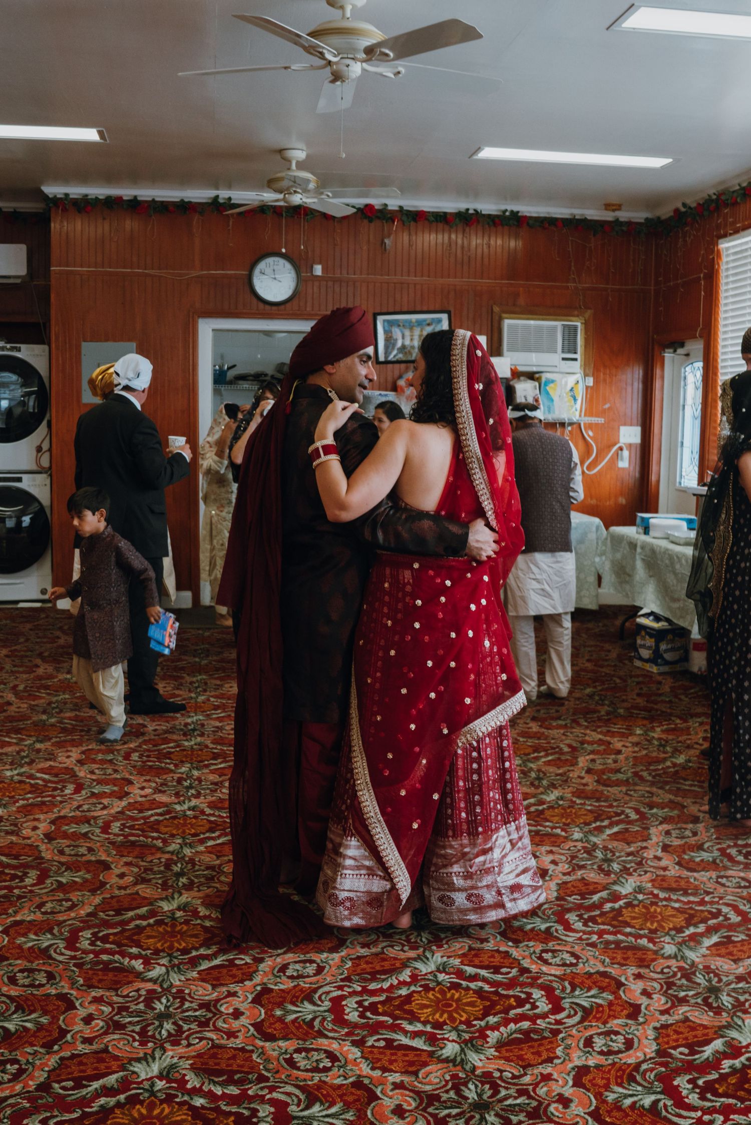Two women in vibrant red Indian wedding attire sharing an emotional embrace at a festive celebration.