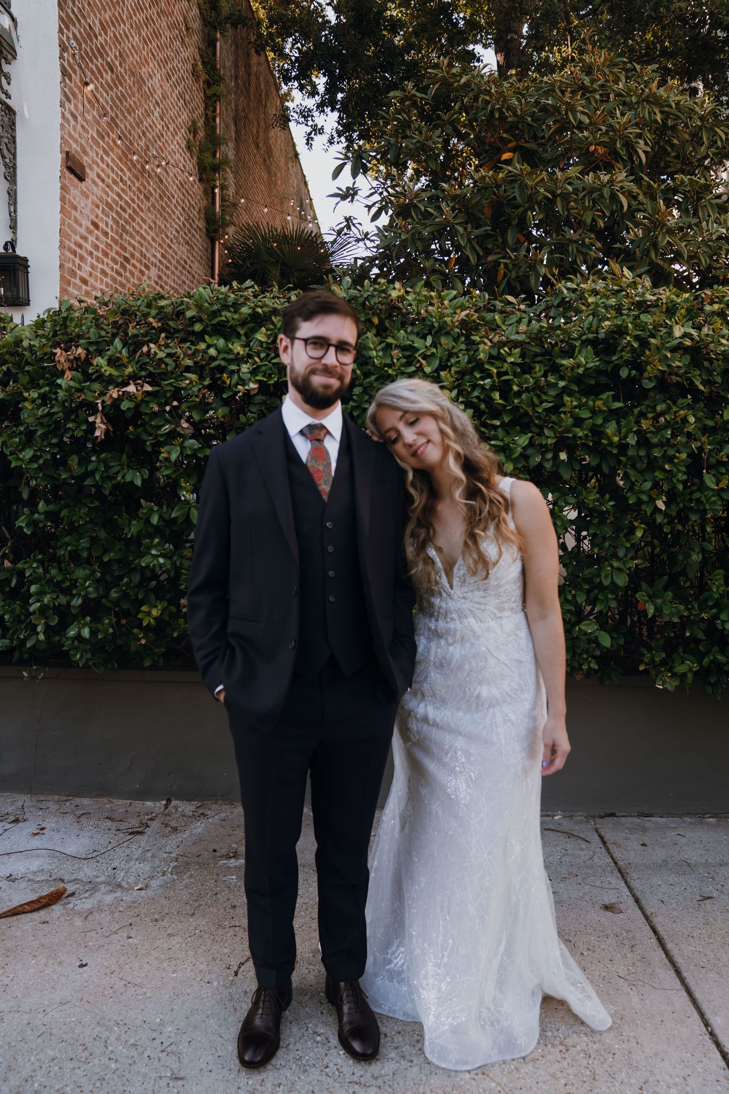 Smiling bride and groom pose together outdoors against a lush green hedge backdrop.