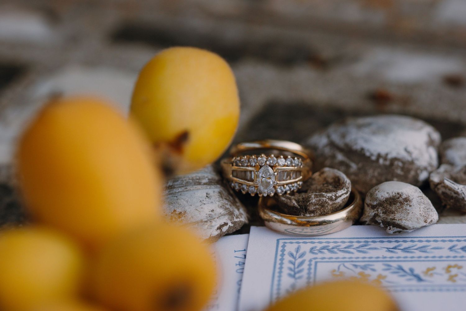 Gold diamond engagement ring and wedding bands rest on stones beside yellow loquat fruits.