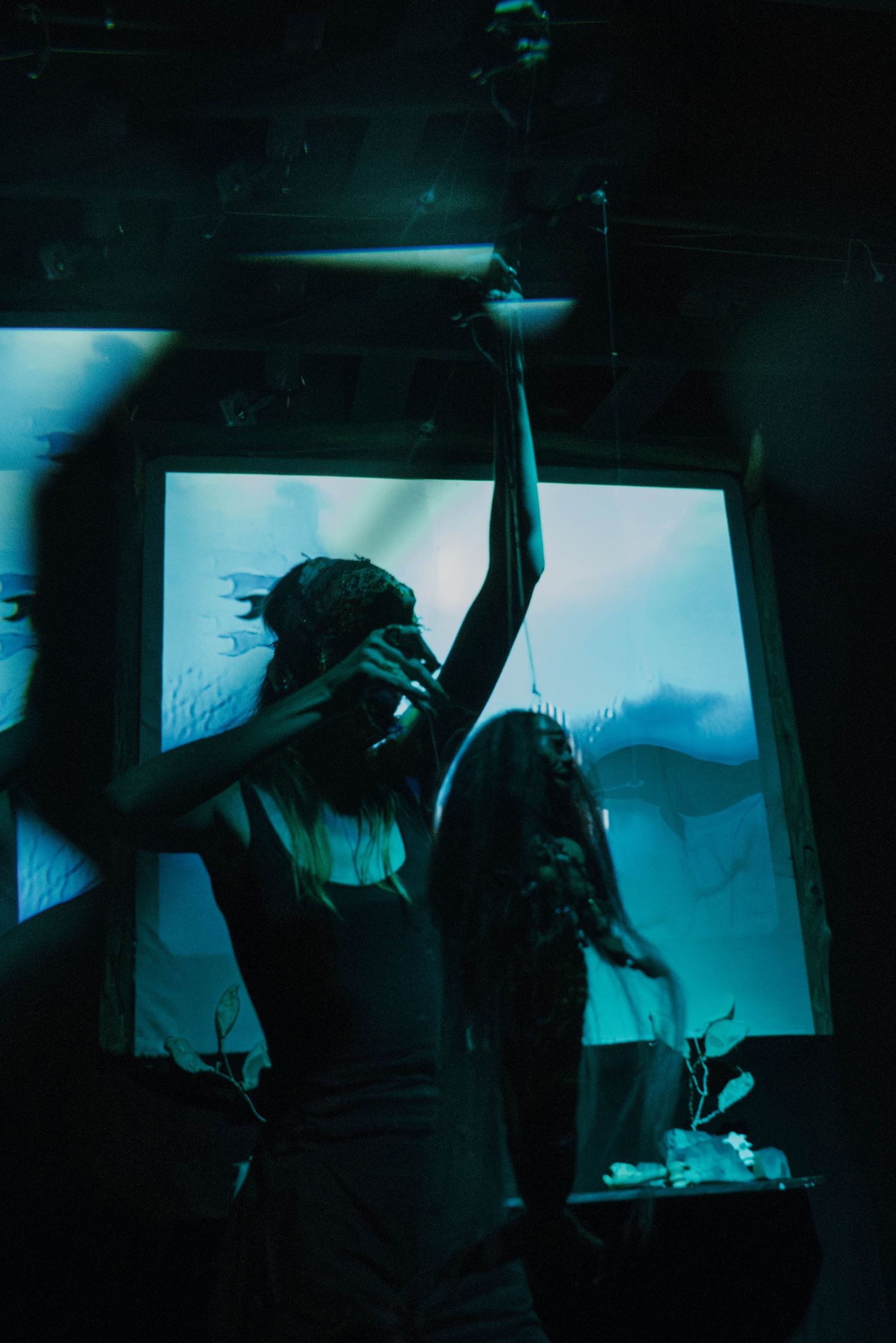 Performer in gas mask reaches upward in teal-lit theatrical scene with projection backdrop.