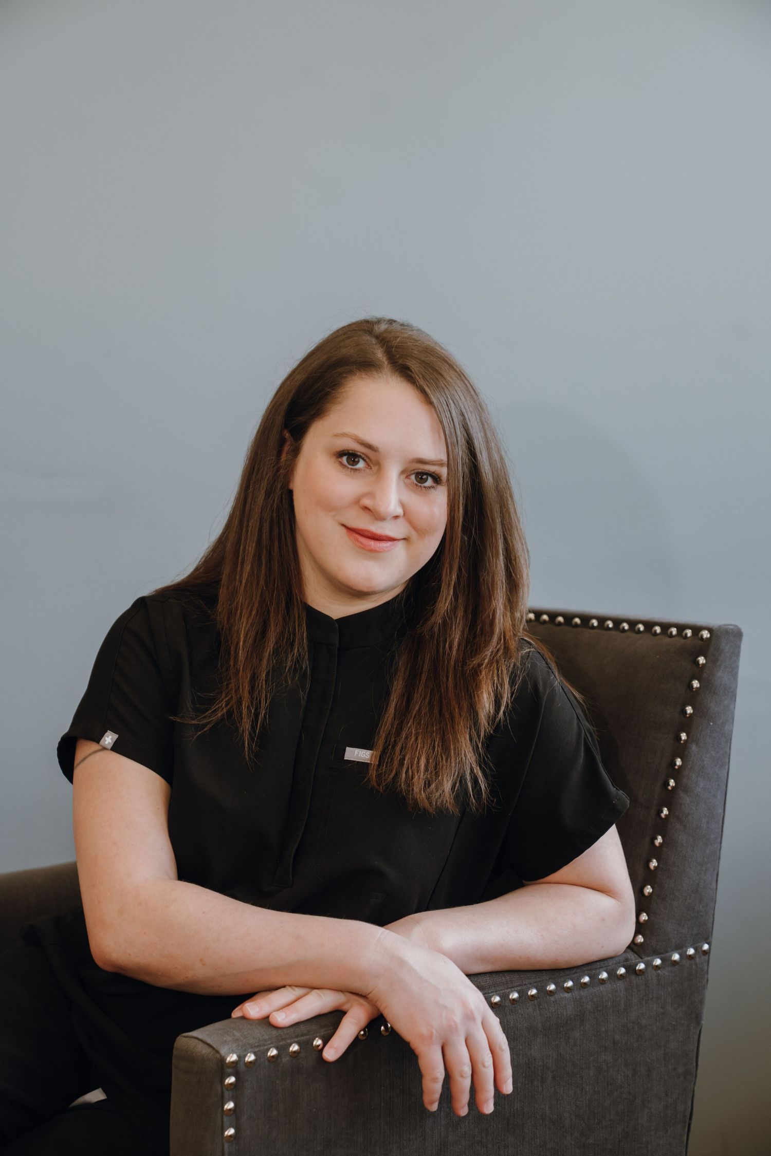 A smiling woman with long brown hair in a black uniform sits relaxed in a studded gray chair.