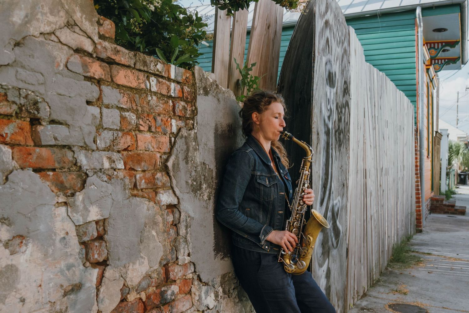 Female musician playing saxophone while leaning against rustic brick wall in narrow alleyway.