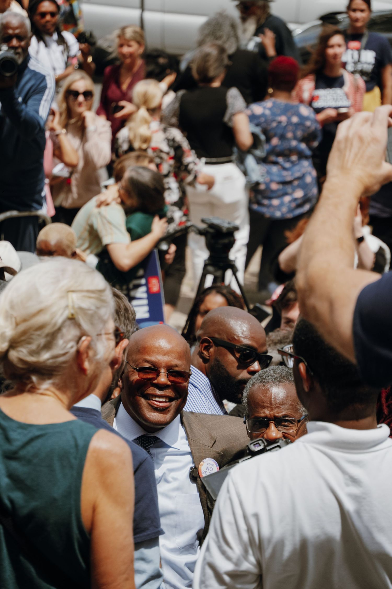 Smiling man in sunglasses and suit surrounded by a diverse crowd at an outdoor political gathering.