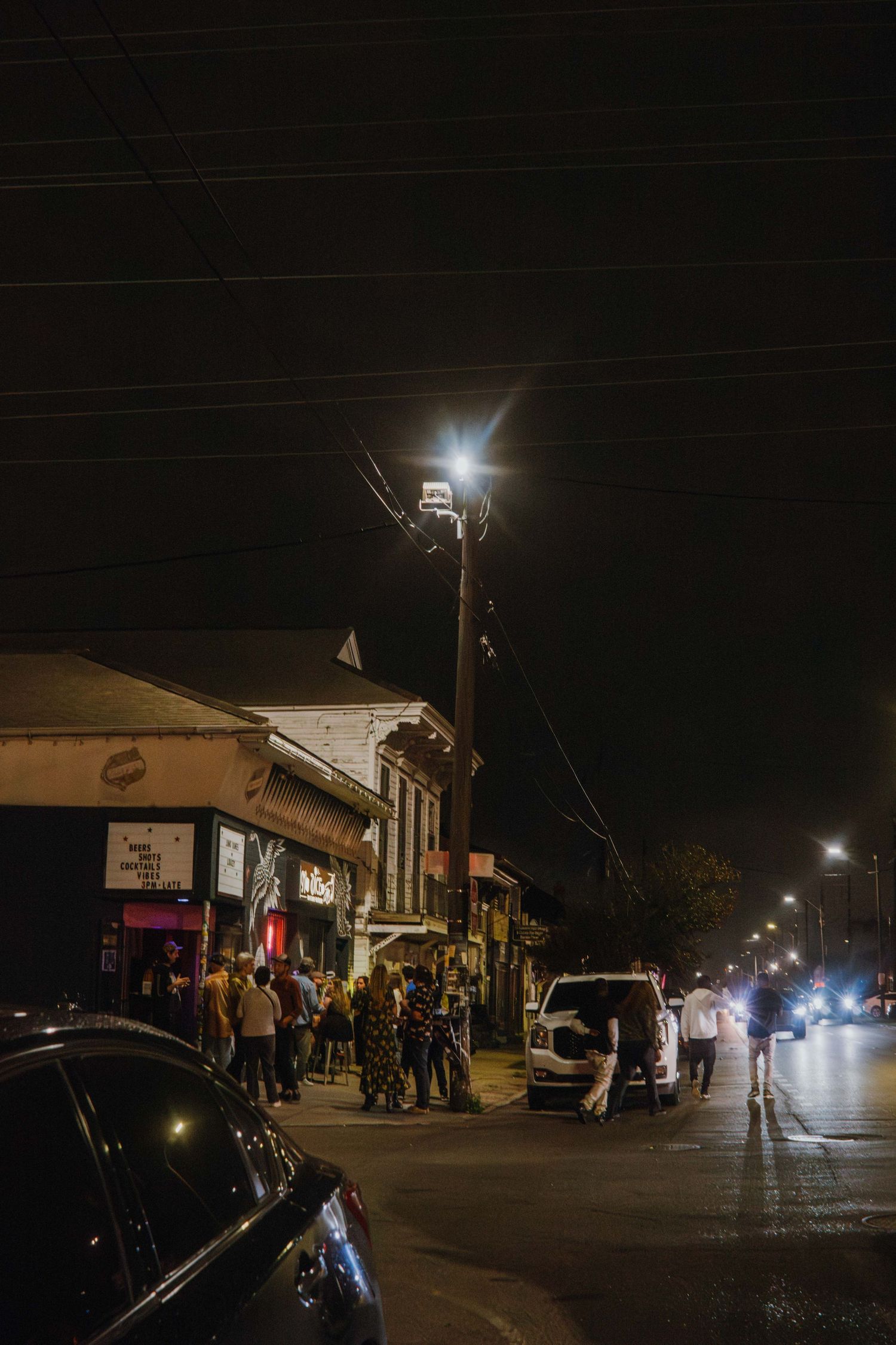 Nighttime street scene with people gathered outside lit storefronts along a busy urban strip.