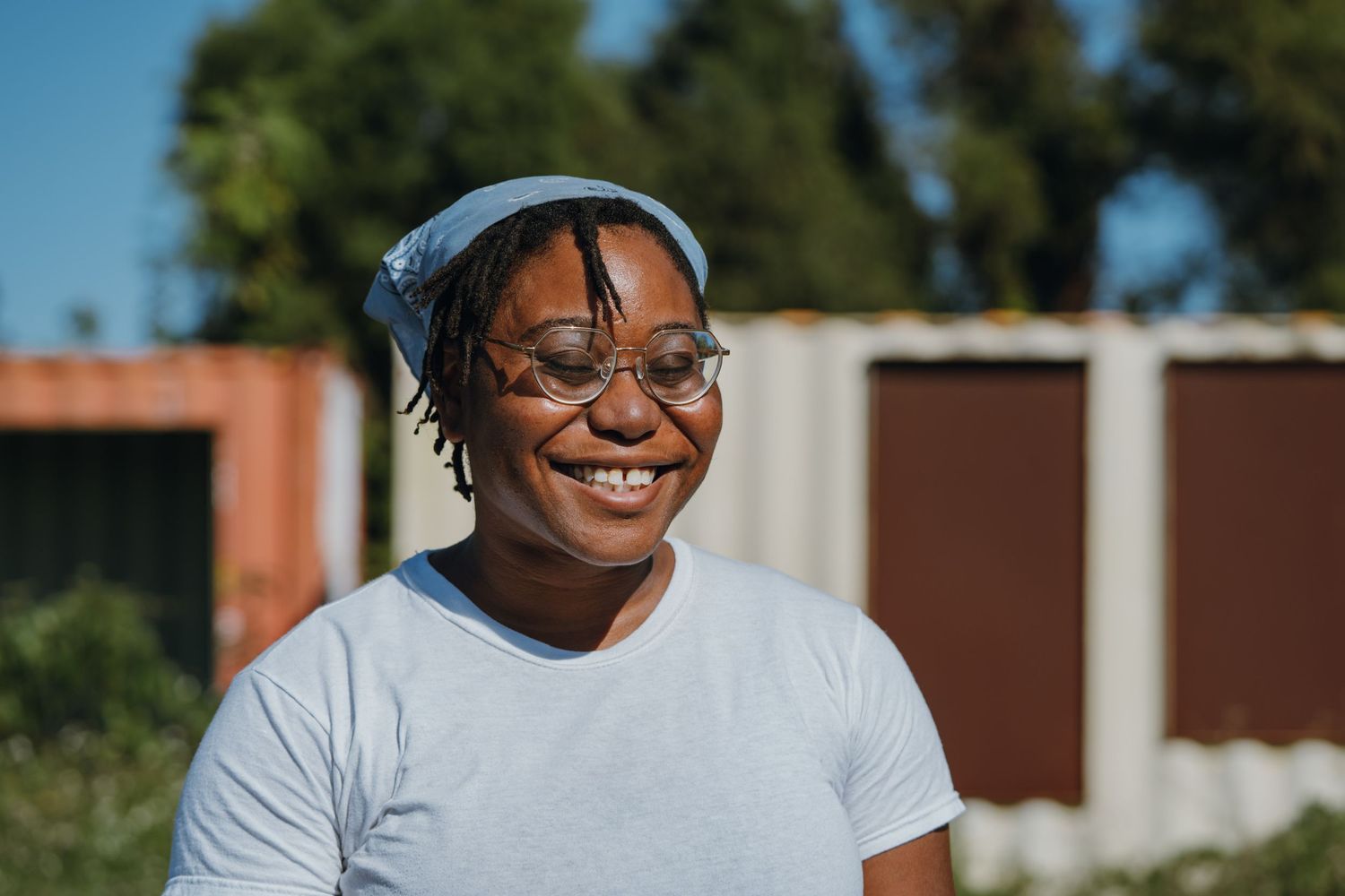 Happy young woman wearing glasses and a blue hat smiling outdoors on a sunny day.