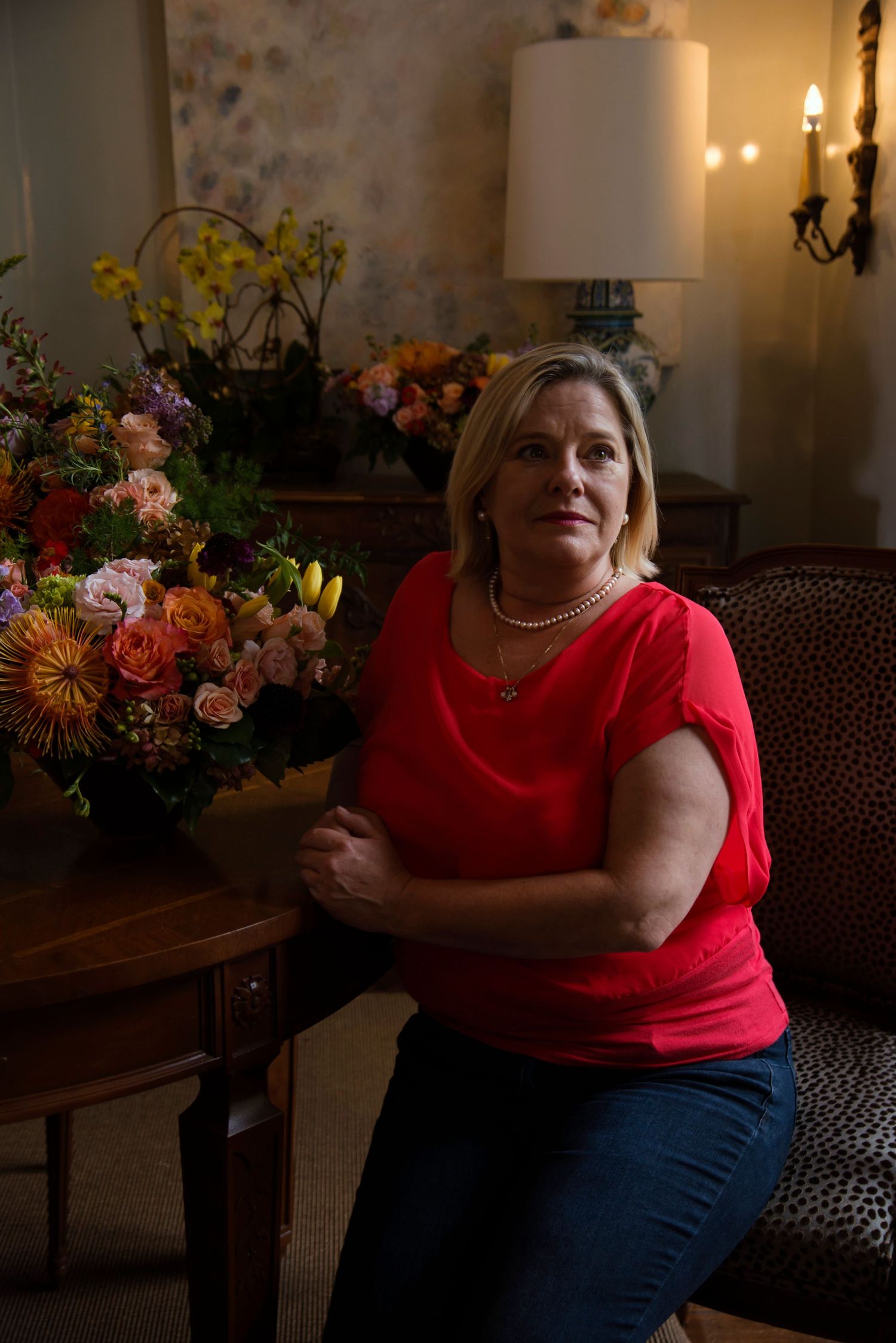Blonde woman in red top sitting beside a colorful floral arrangement in a warmly lit room.
