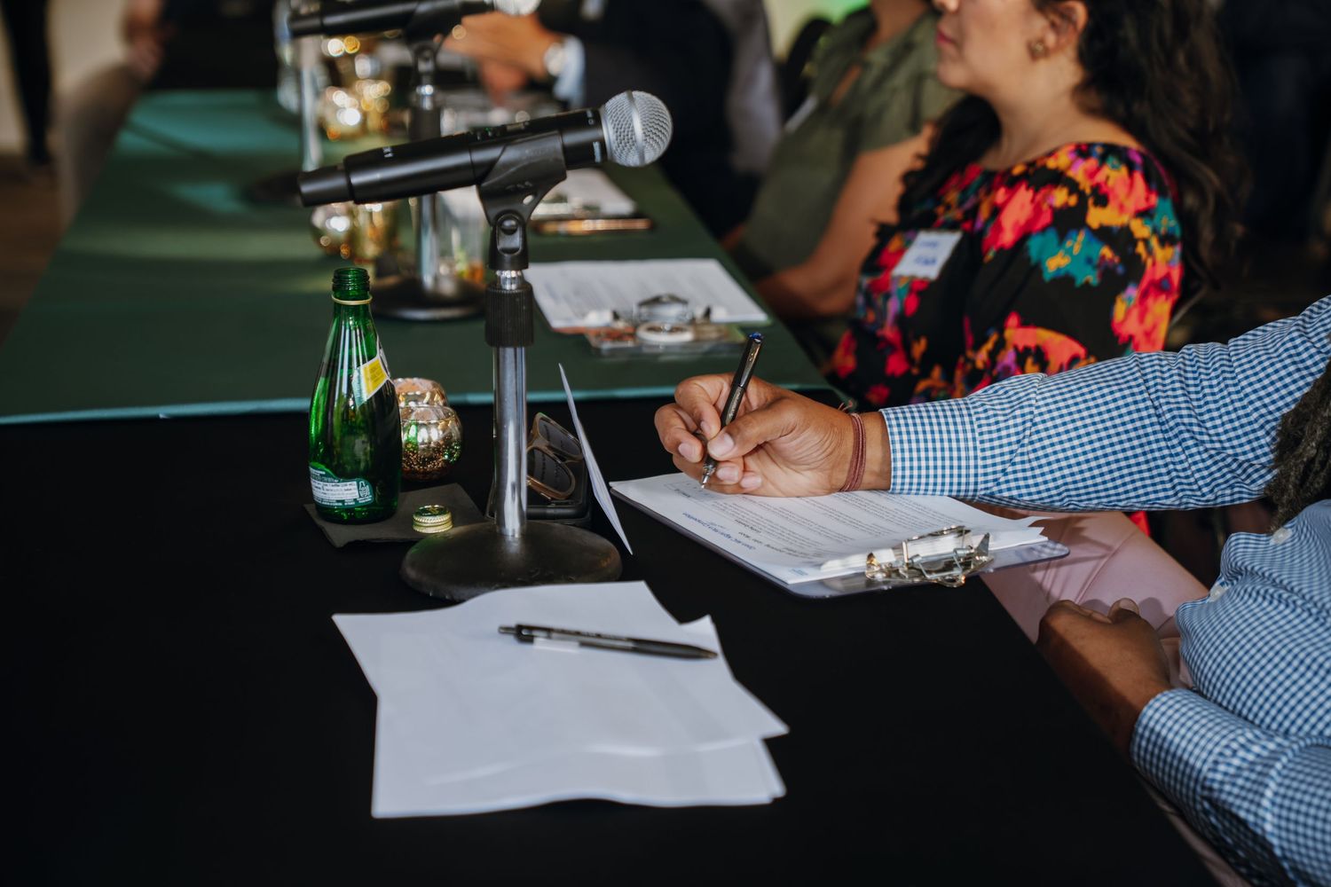 Person writes on clipboard at conference table with microphone and water bottles.