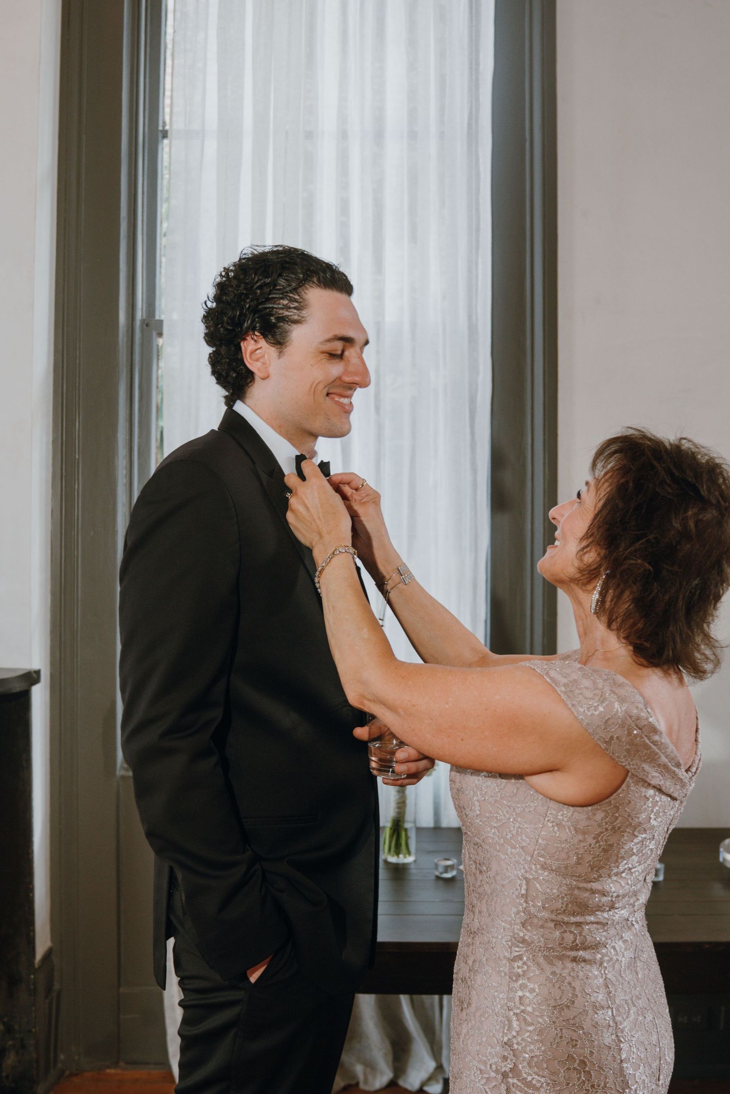 Smiling groom in black tuxedo having his bow tie adjusted by his mother before the wedding ceremony.