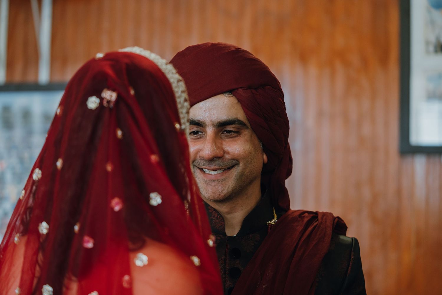 Smiling Indian groom in dark sherwani and maroon turban embracing bride in red wedding veil.