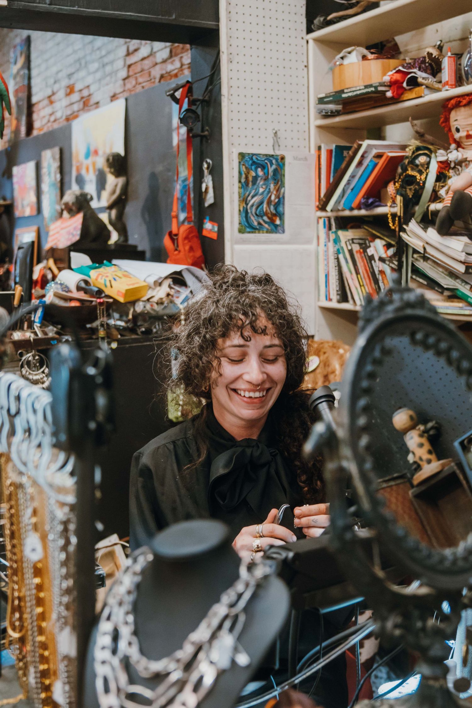 Smiling woman with curly hair browsing jewelry in a cluttered vintage shop filled with eclectic items.