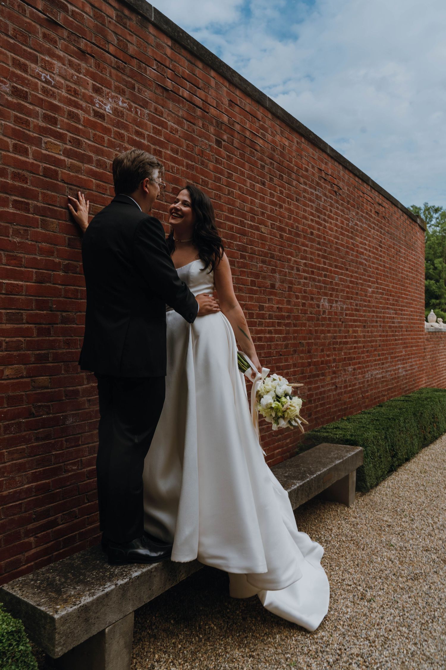 Laughing bride in white gown holds bouquet while embracing groom against a rustic red brick wall outdoors. New Orleans, Louisiana, Mississip